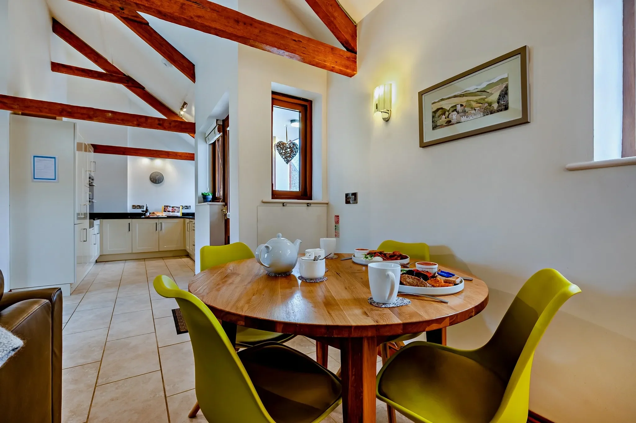 Barley Park dining area with a round oak table and green chairs set for breakfast, looking through to the fitted kitchen beyond.