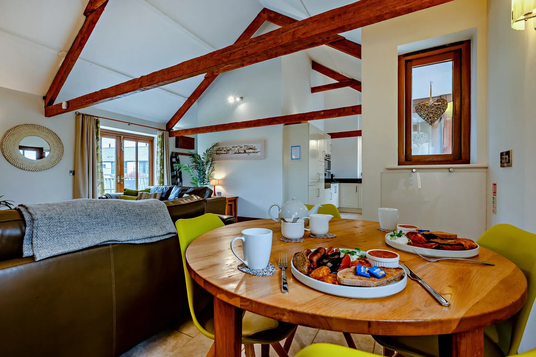 Barley Park dining table with green chairs in the foreground, with the lounge seating area and kitchen visible beyond under vaulted beams.