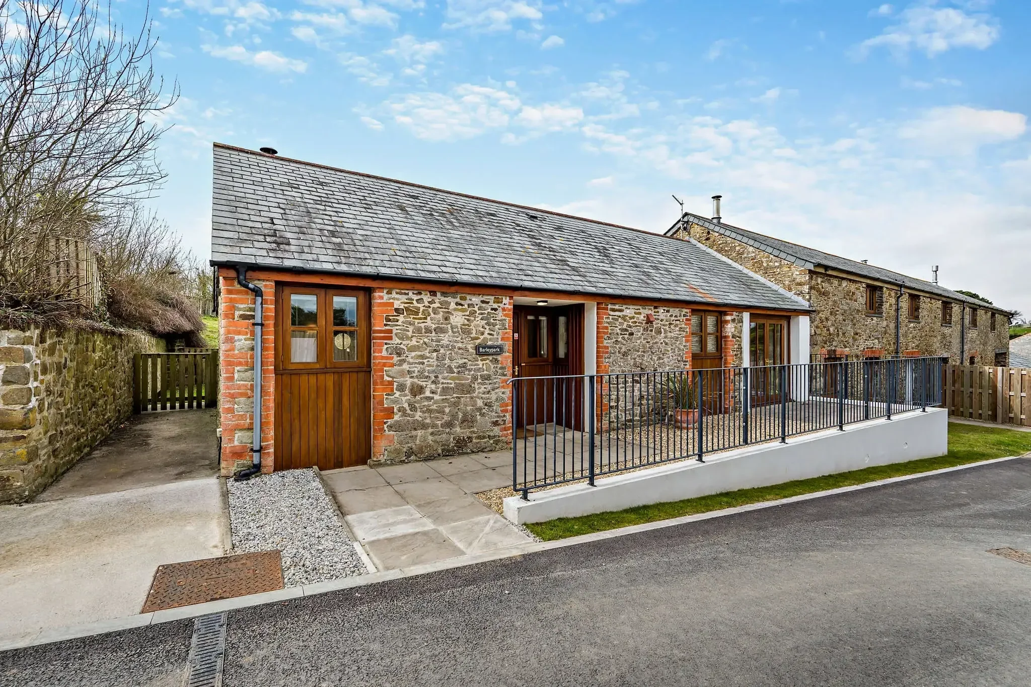 Barley Park stone-built cottage seen from the road with slate roof, timber-framed windows, iron railings and a garage to the left.