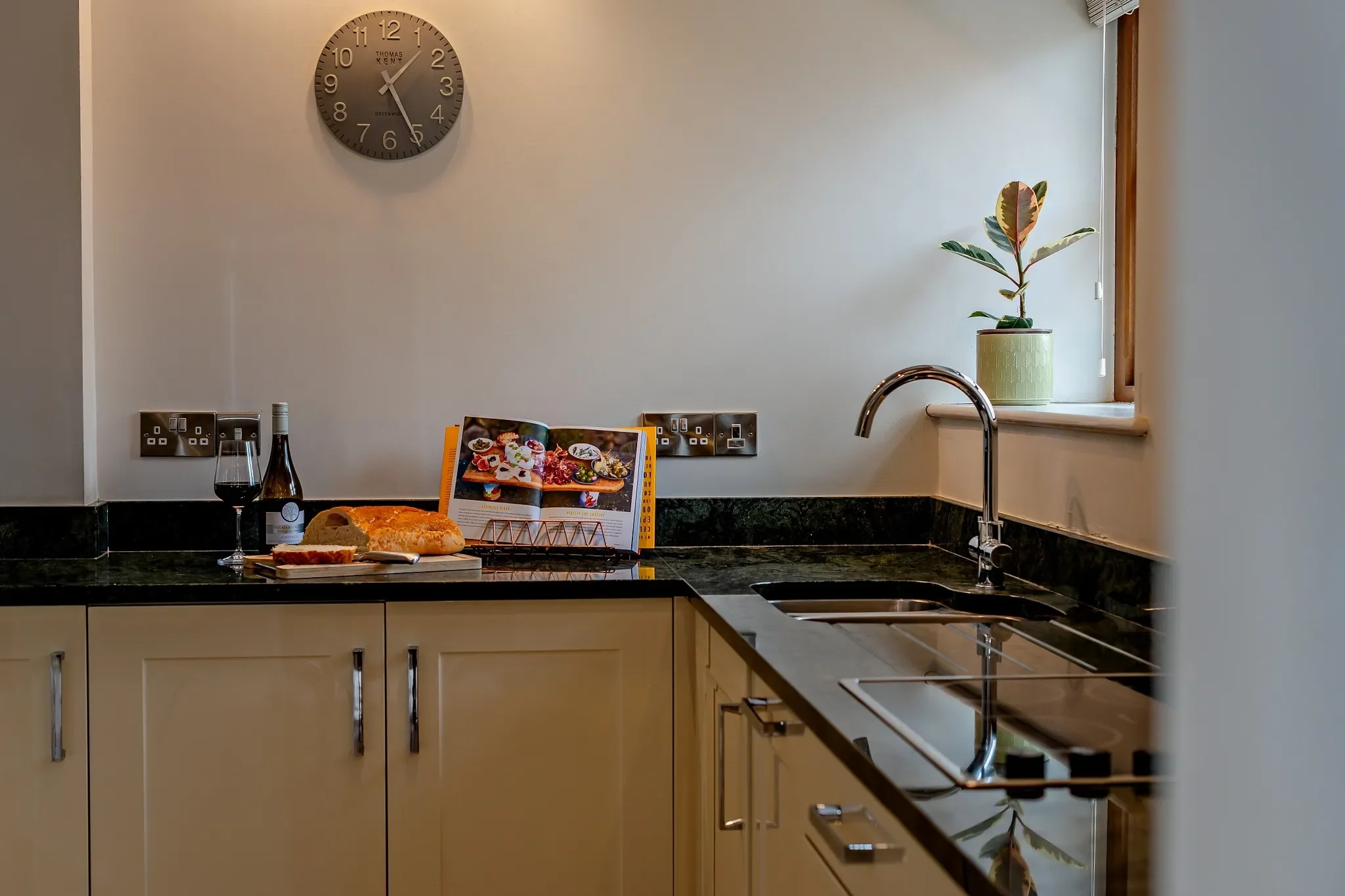 Barley Park kitchen worktop and sink detail showing dark granite counters, chrome tap and a cookbook propped by a window.