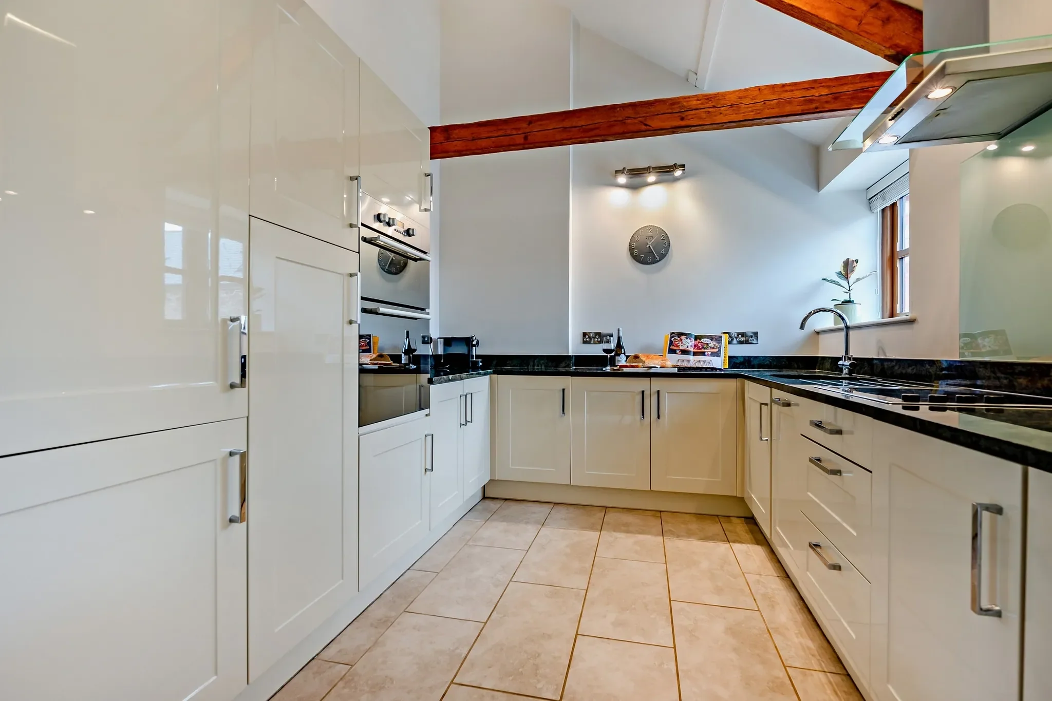 Barley Park kitchen with gloss white cabinetry, dark granite worktops, double oven and exposed ceiling beam in a vaulted space.