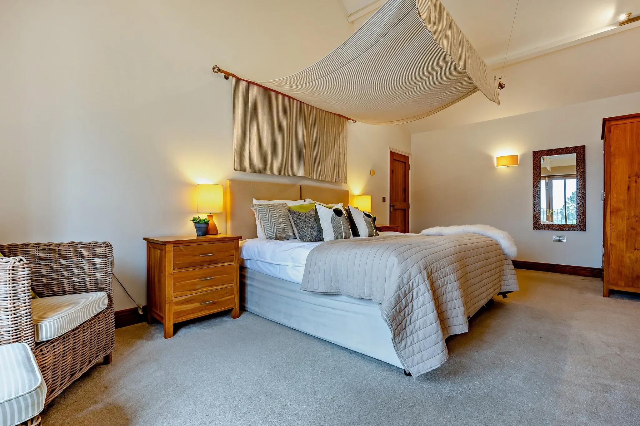 Barley Park master bedroom with a draped four-poster bed, wicker armchair and warm bedside lighting under a vaulted ceiling.