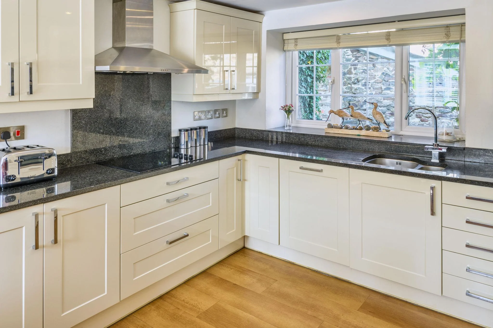 Calf House kitchen with cream shaker cabinets, dark granite worktops, stainless-steel range hood, and a window overlooking the stone wall outside.