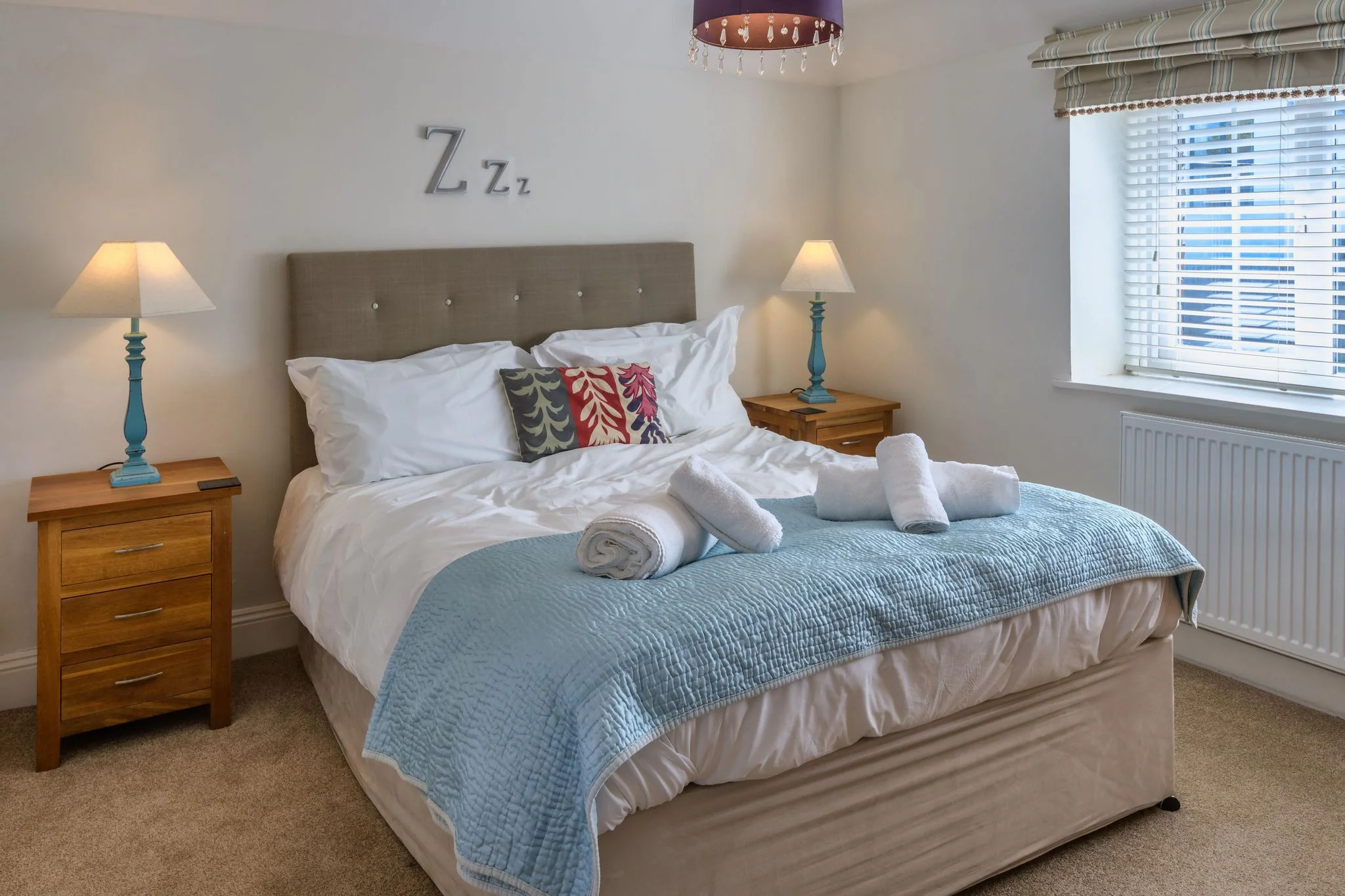Calf House master bedroom with upholstered double bed, teal bedside lamps, and fresh white towels laid out on a duck-egg throw.