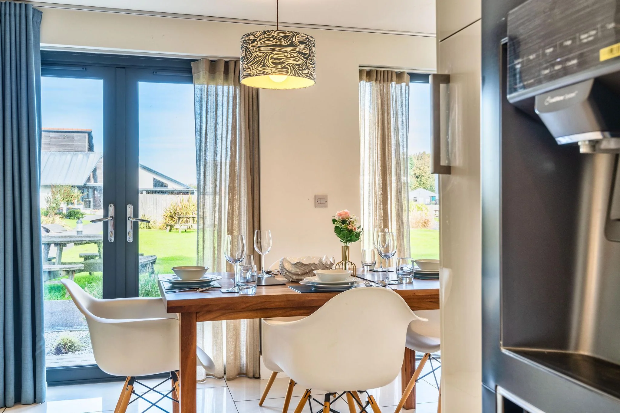 Chapel Park dining area set for six with a walnut table, modern white chairs, and bi-fold doors framing a sunny garden view.
