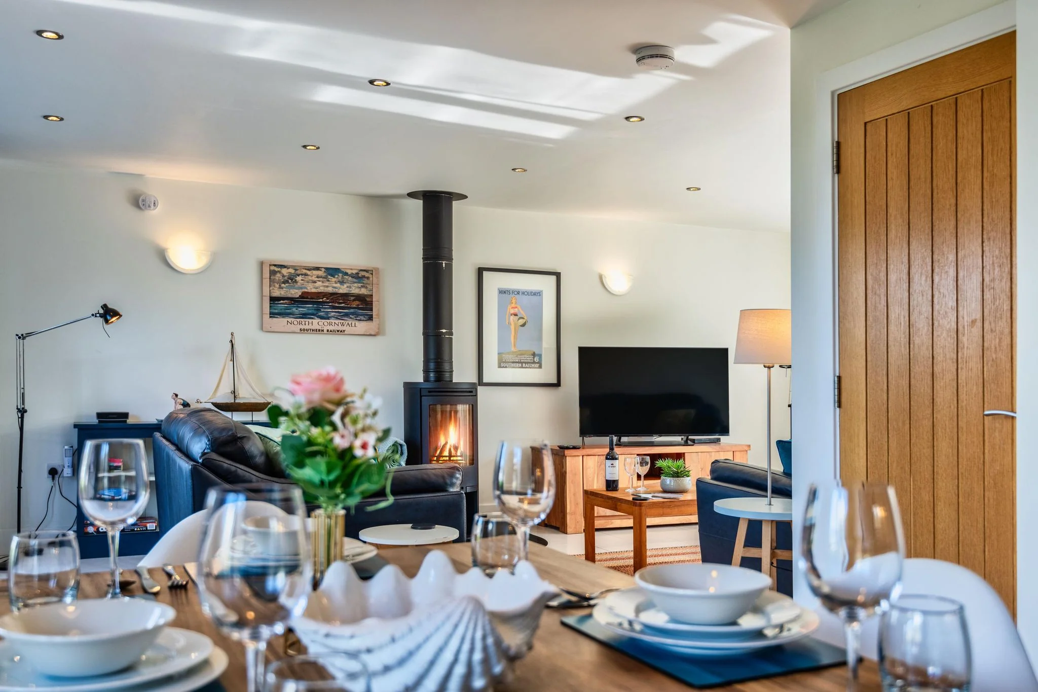 Chapel Park dining table dressed for dinner in the foreground, looking through to the sitting room with its glowing wood burner and coastal artwork beyond.