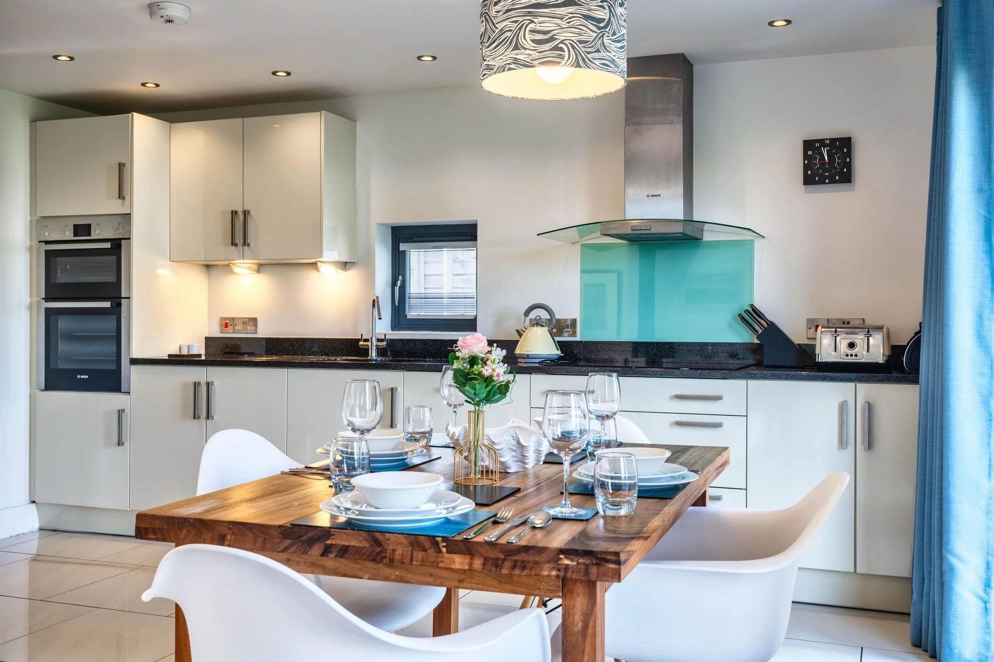 Chapel Park kitchen with white gloss cabinetry, teal splashback, double oven, and the dining table set with glassware in the foreground.