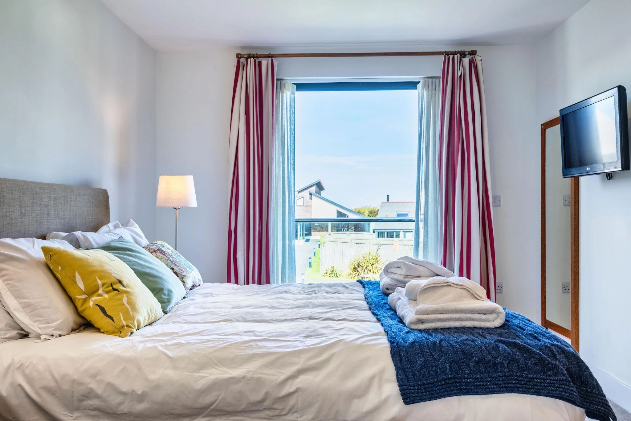 Chapel Park master bedroom with a king-size bed, red-striped curtains, wall-mounted TV, and a Juliet balcony window with views over neighbouring rooftops.