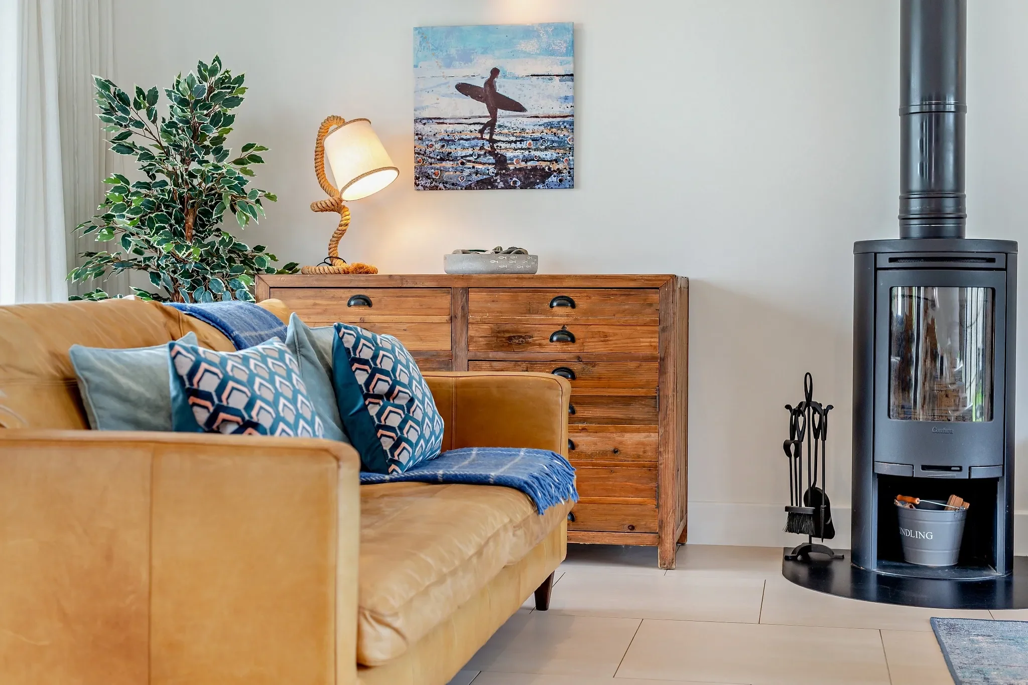 Eagles Nest sitting area corner showing the freestanding wood-burning stove, mustard sofa and coastal surf artwork.
