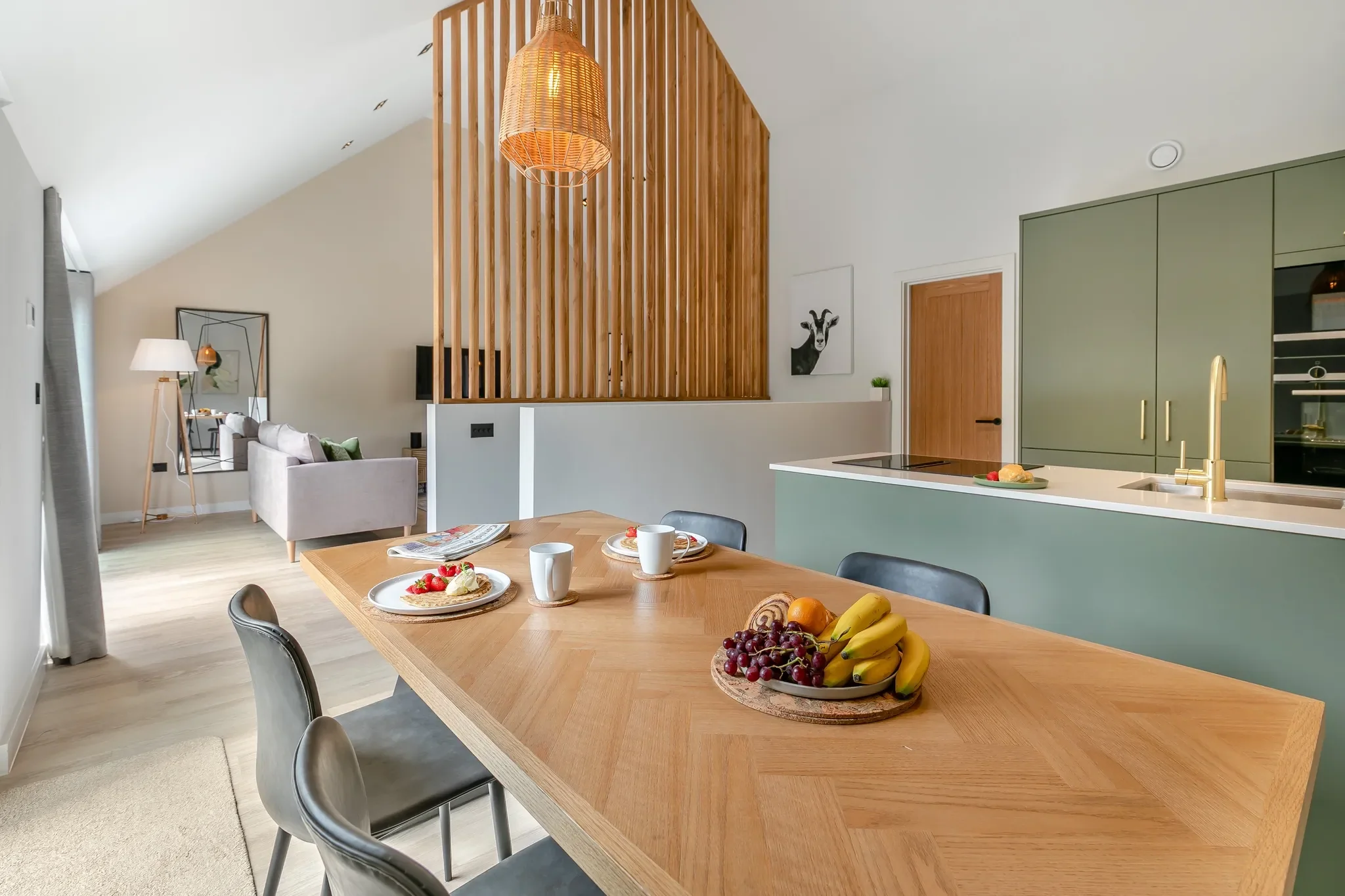 Gwari Spa Barn open-plan dining and kitchen with herringbone oak table, sage-green cabinetry and island beyond the slatted screen.