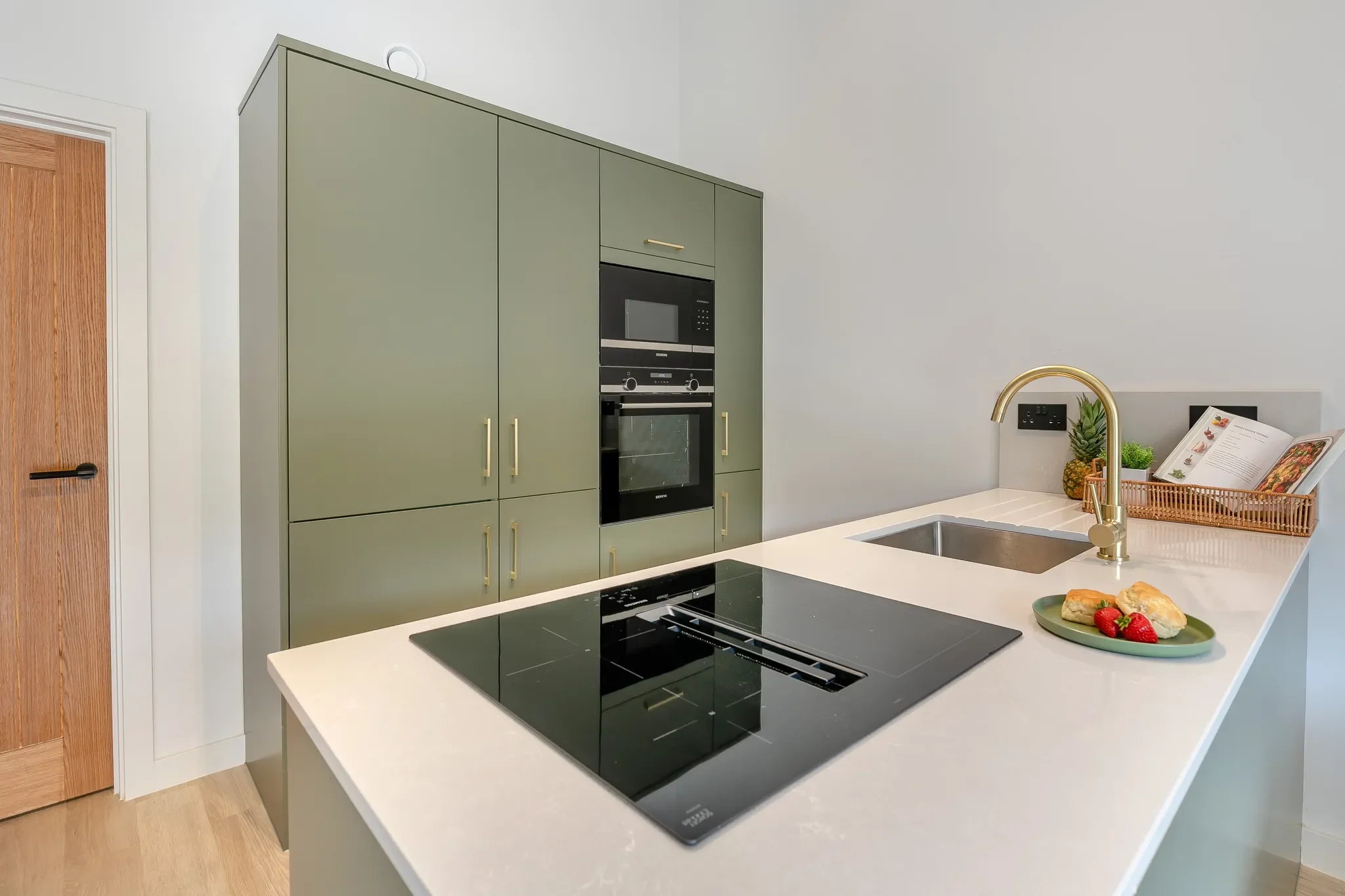 Gwari Spa Barn sage-green kitchen island with induction hob, integrated oven and brass tap on a quartz worktop.