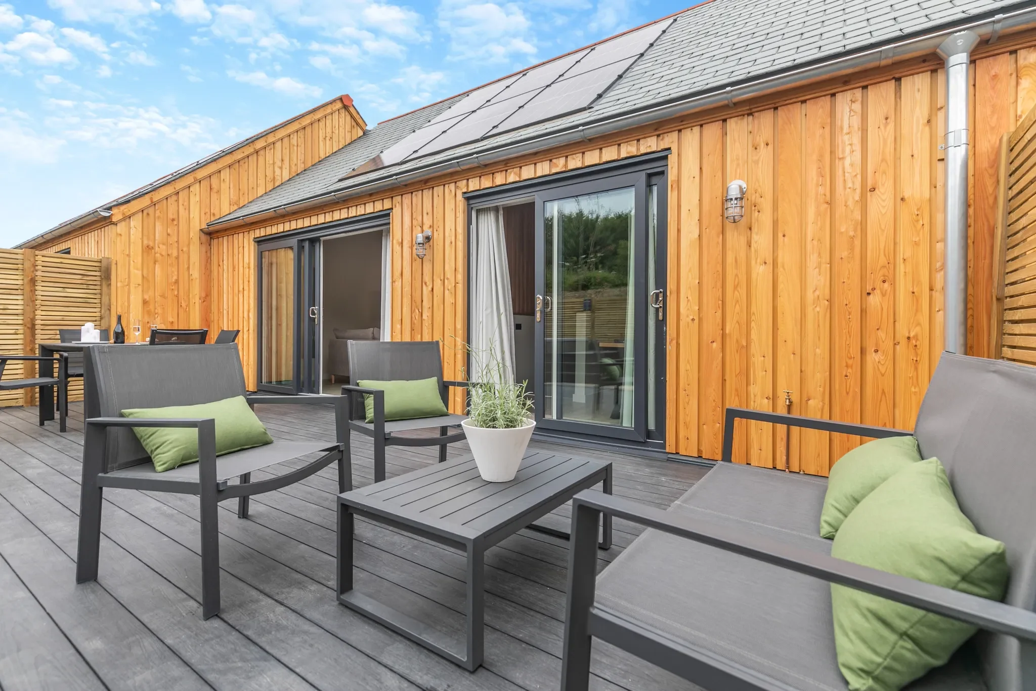 Gwari Spa Barn upper deck terrace with grey outdoor lounge furniture, green cushions and solar-panelled barn roof behind.