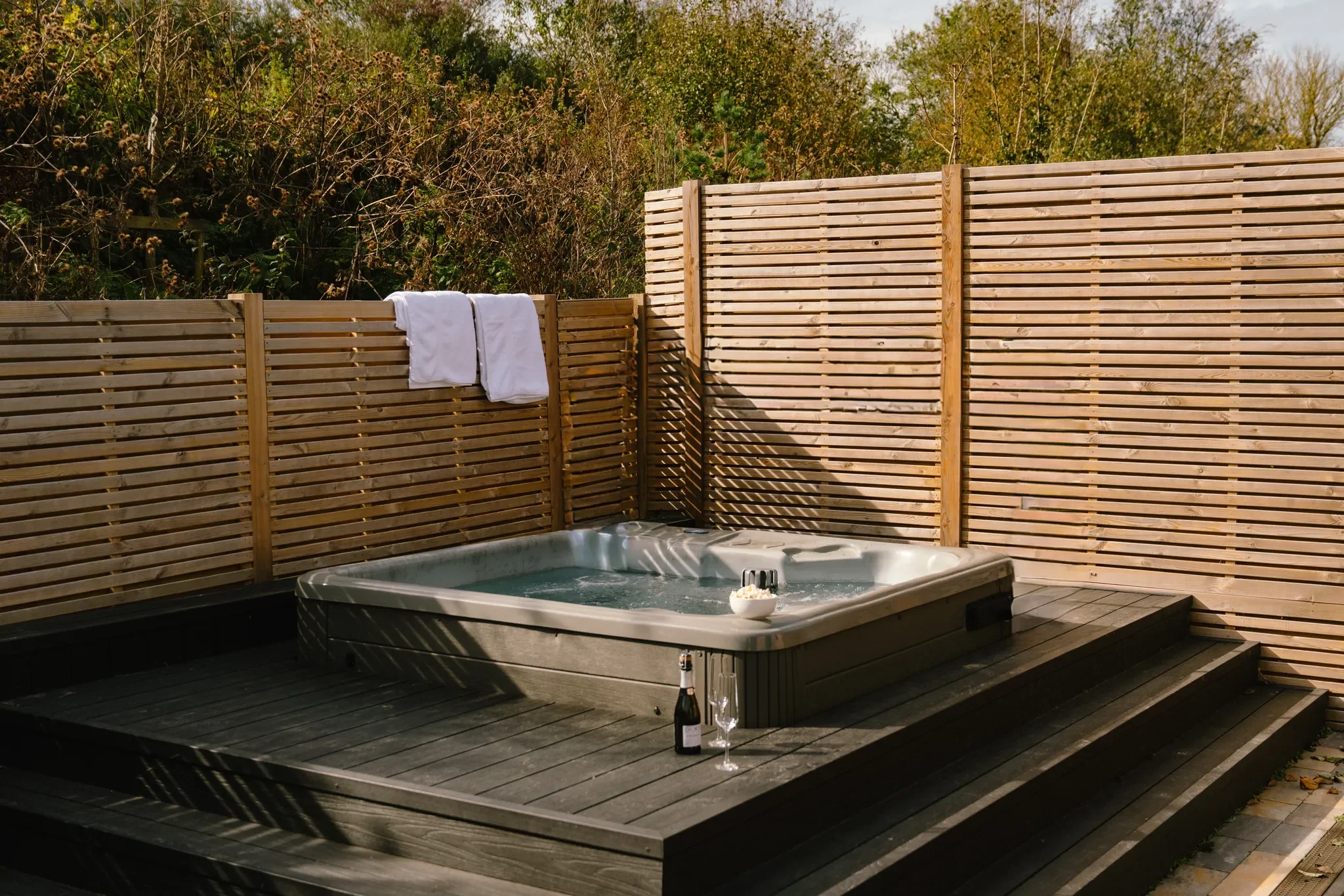 Gwari Spa Barn private hot tub on a hardwood deck, enclosed by slatted timber screens with towels and champagne set out.
