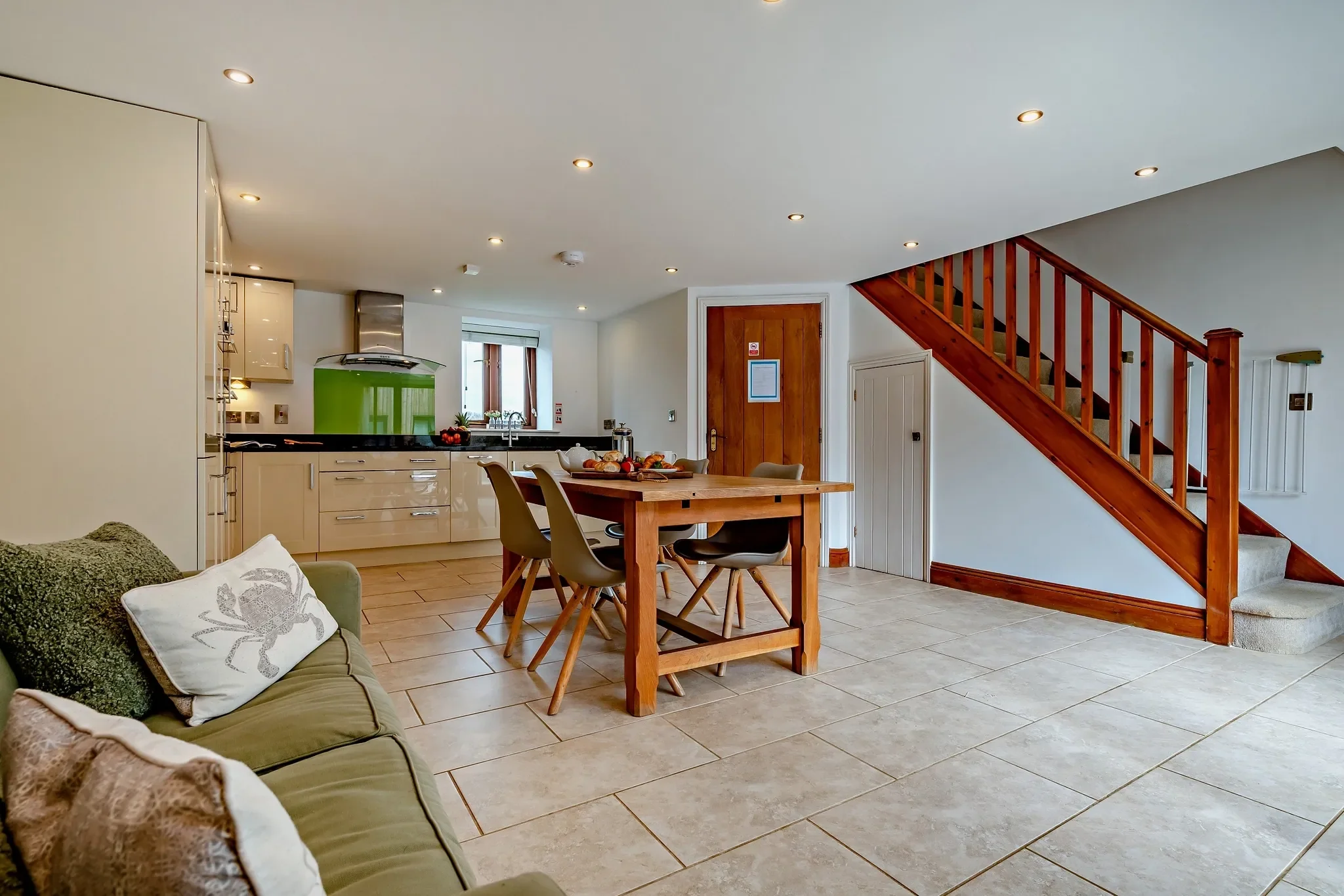 Jack's House kitchen-diner showing the exposed timber staircase rising alongside the green-splashback kitchen and dining table.