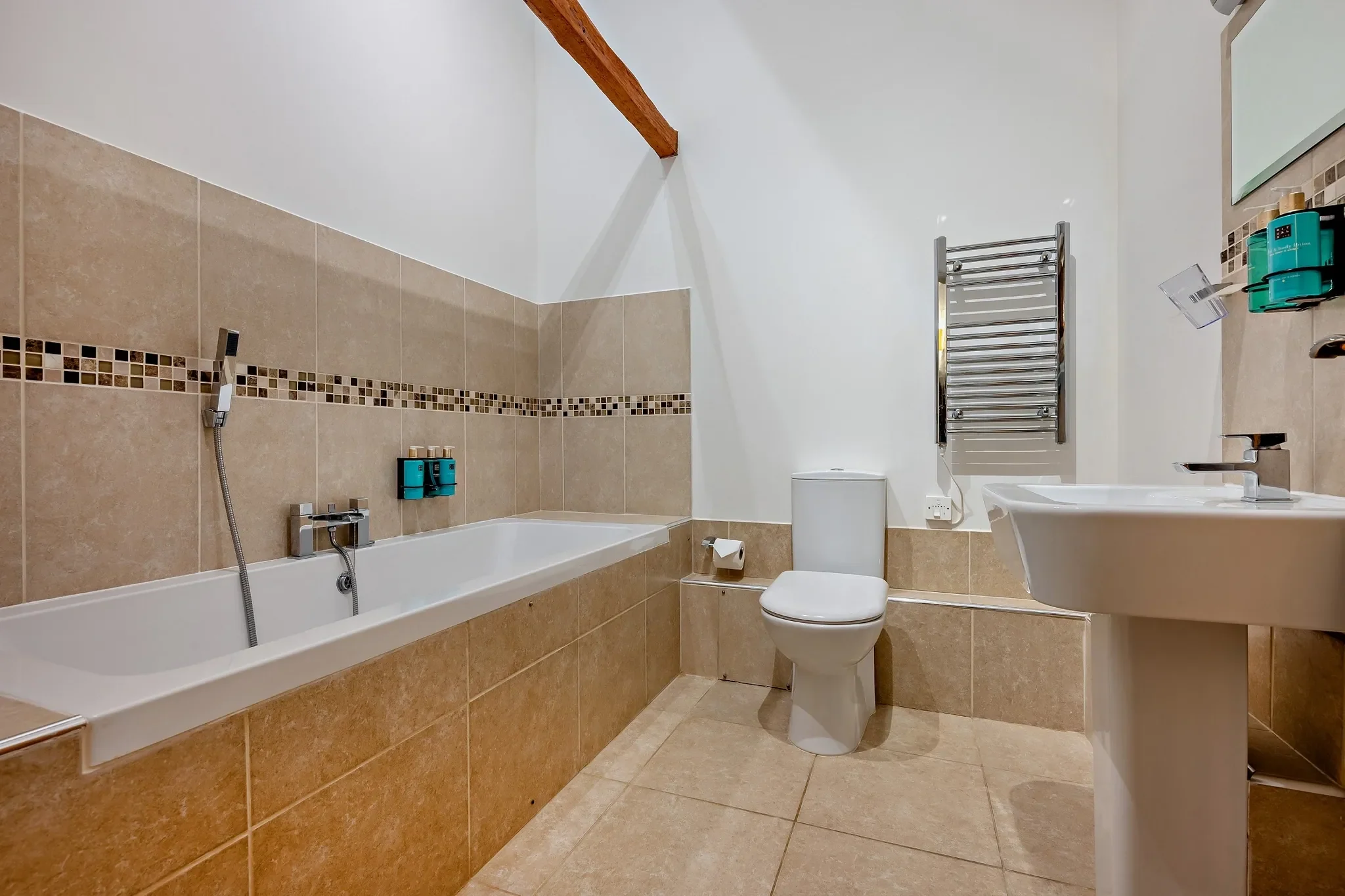 Long Down bathroom with full-size bath, pedestal basin, heated towel rail and mosaic tile border under a vaulted beam.