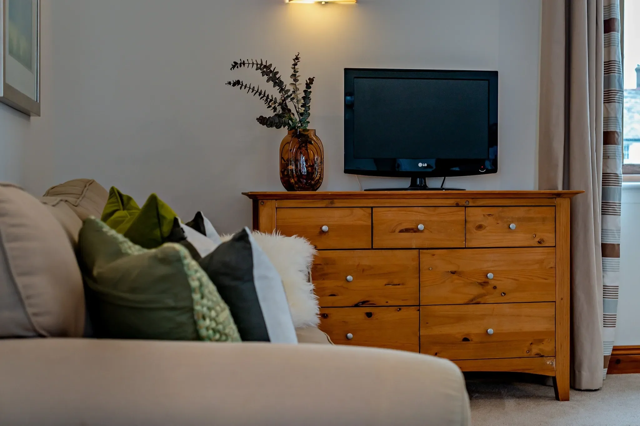 Long Down master bedroom detail of pine chest of drawers with TV and eucalyptus vase beside the sofa corner.