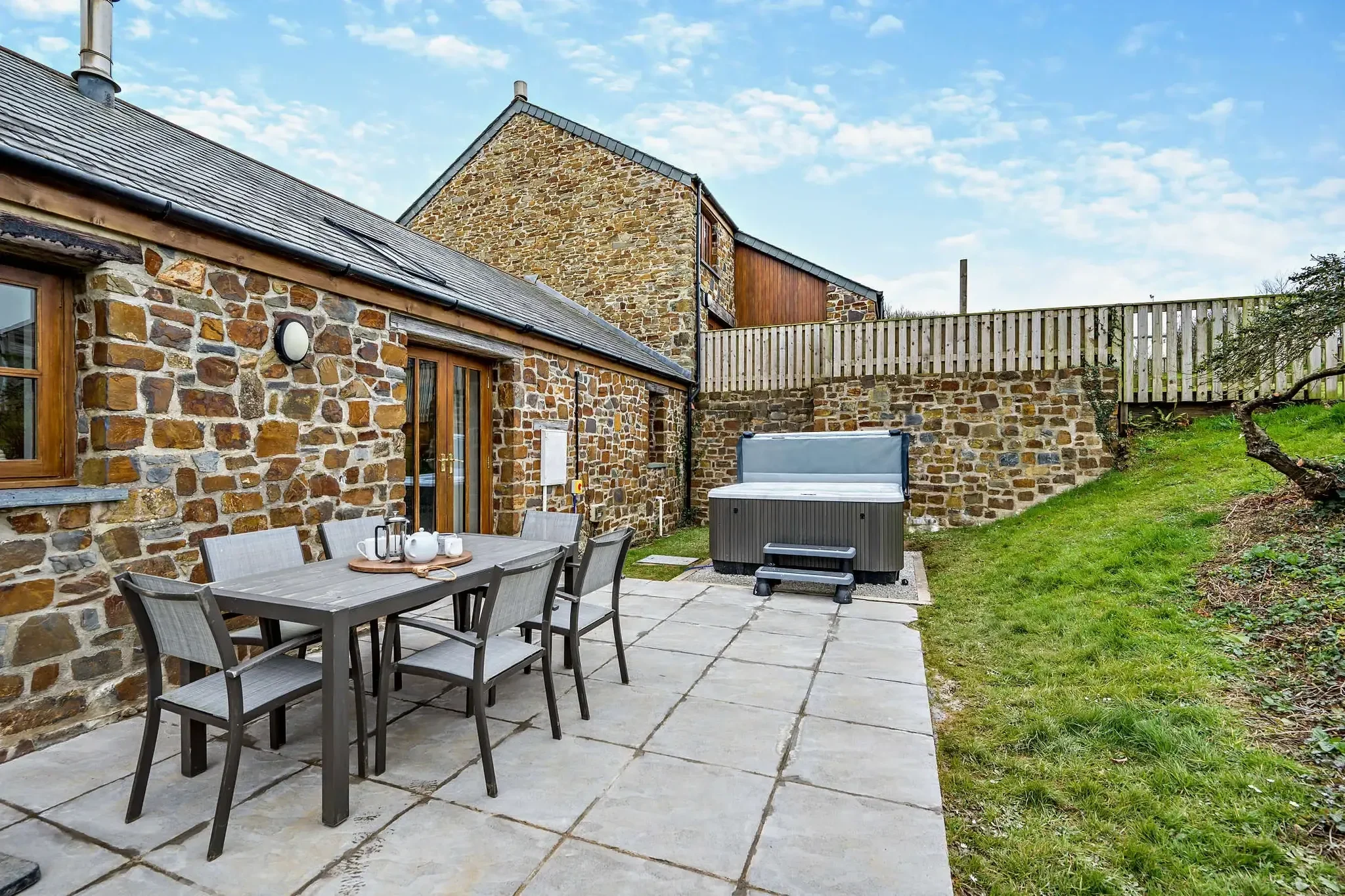 Long Down rear stone terrace with six-seat dining set and the hot tub alongside the cottage wall under open sky.