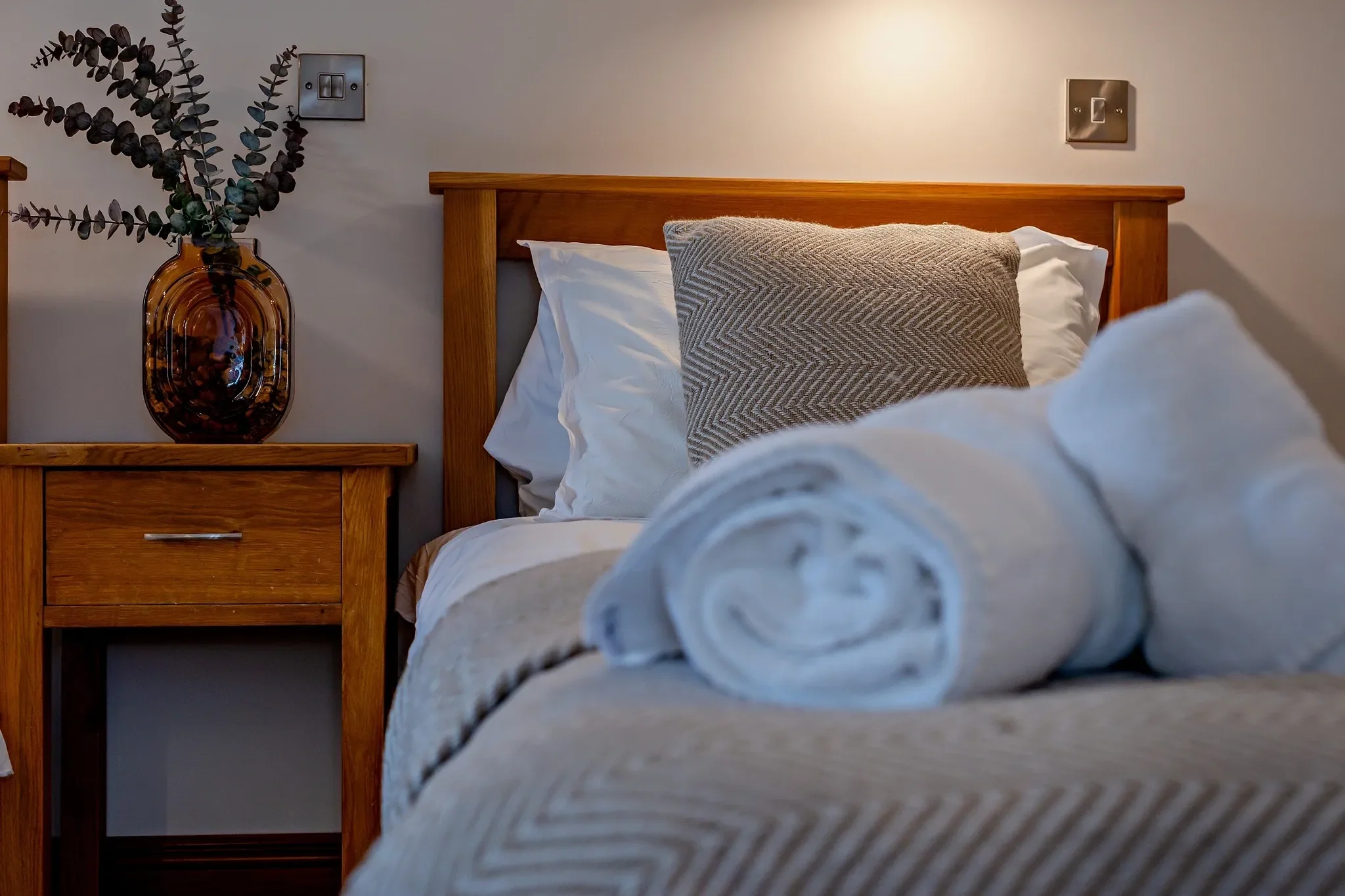 Long Down twin room close-up of a single bed with rolled towels on the herringbone bedspread and amber vase on the nightstand.