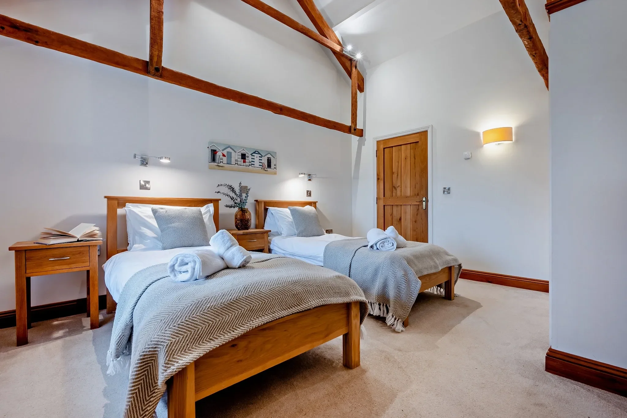 Long Down twin bedroom with two single beds dressed in grey, exposed A-frame beams and coastal beach-hut shelf artwork.