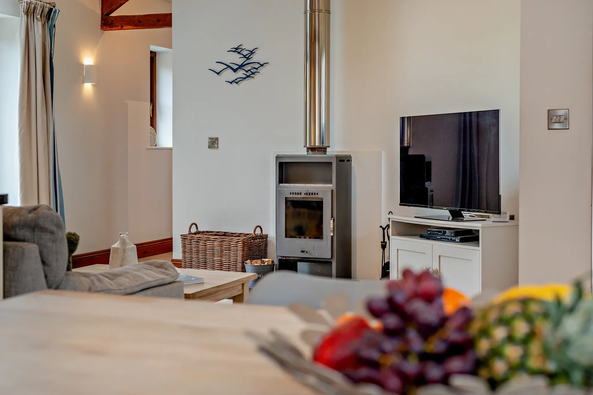 Long Down lounge close-up of the log-burning stove, wicker log basket and flat-screen TV with coastal bird wall art above.