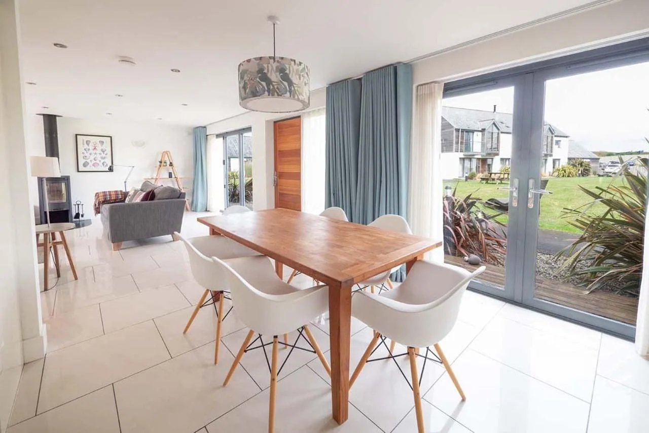 Medlands dining area with six-seat oak table, pendant light and full-width bi-fold doors overlooking the garden and neighbouring cottages.