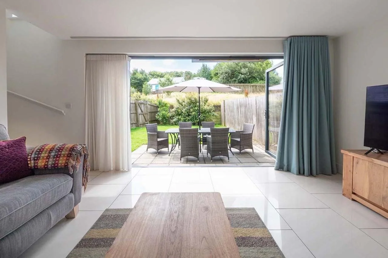 Medlands living room looking through fully open bi-fold doors to the private patio, rattan garden furniture and lawned rear garden.
