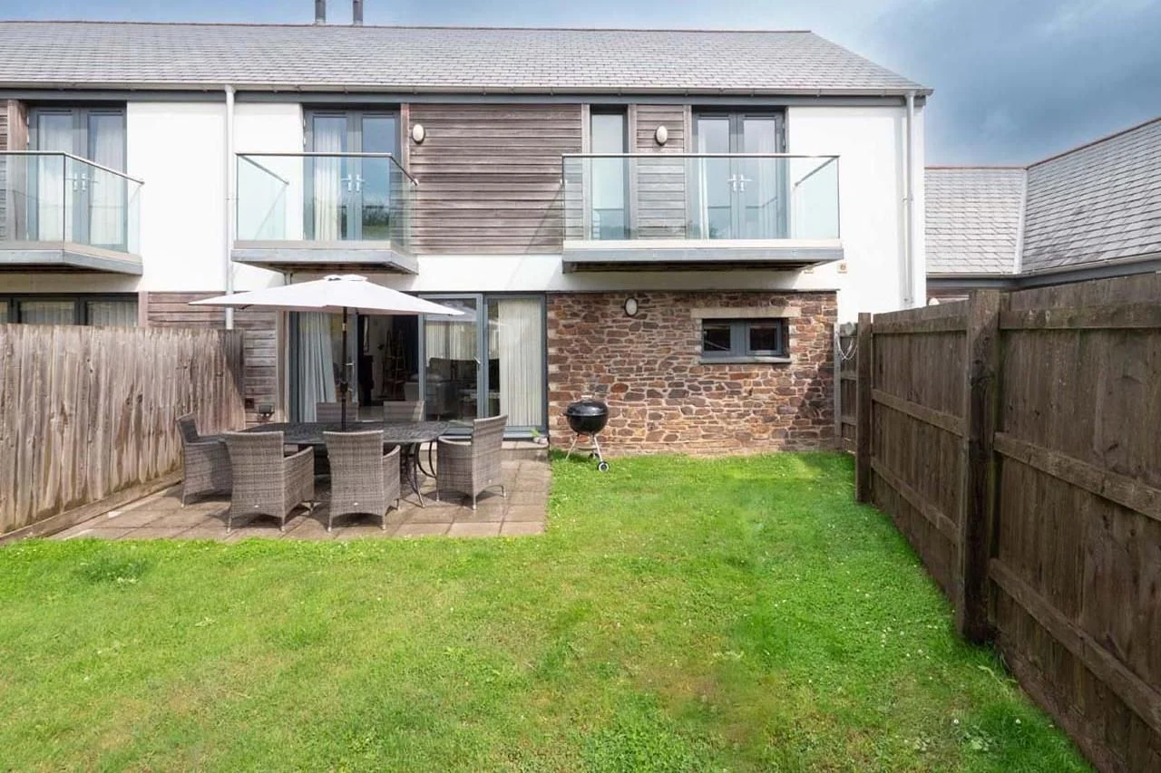 Medlands rear garden with paved patio, rattan dining set and parasol, lawned area and timber balcony above.