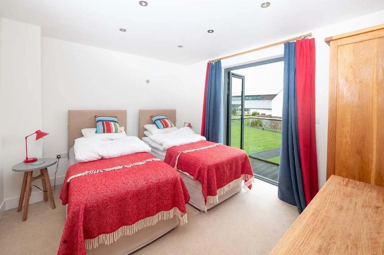 Medlands twin bedroom with two single beds in red throws, blue and red curtains and bi-fold doors looking onto the estate lawn.