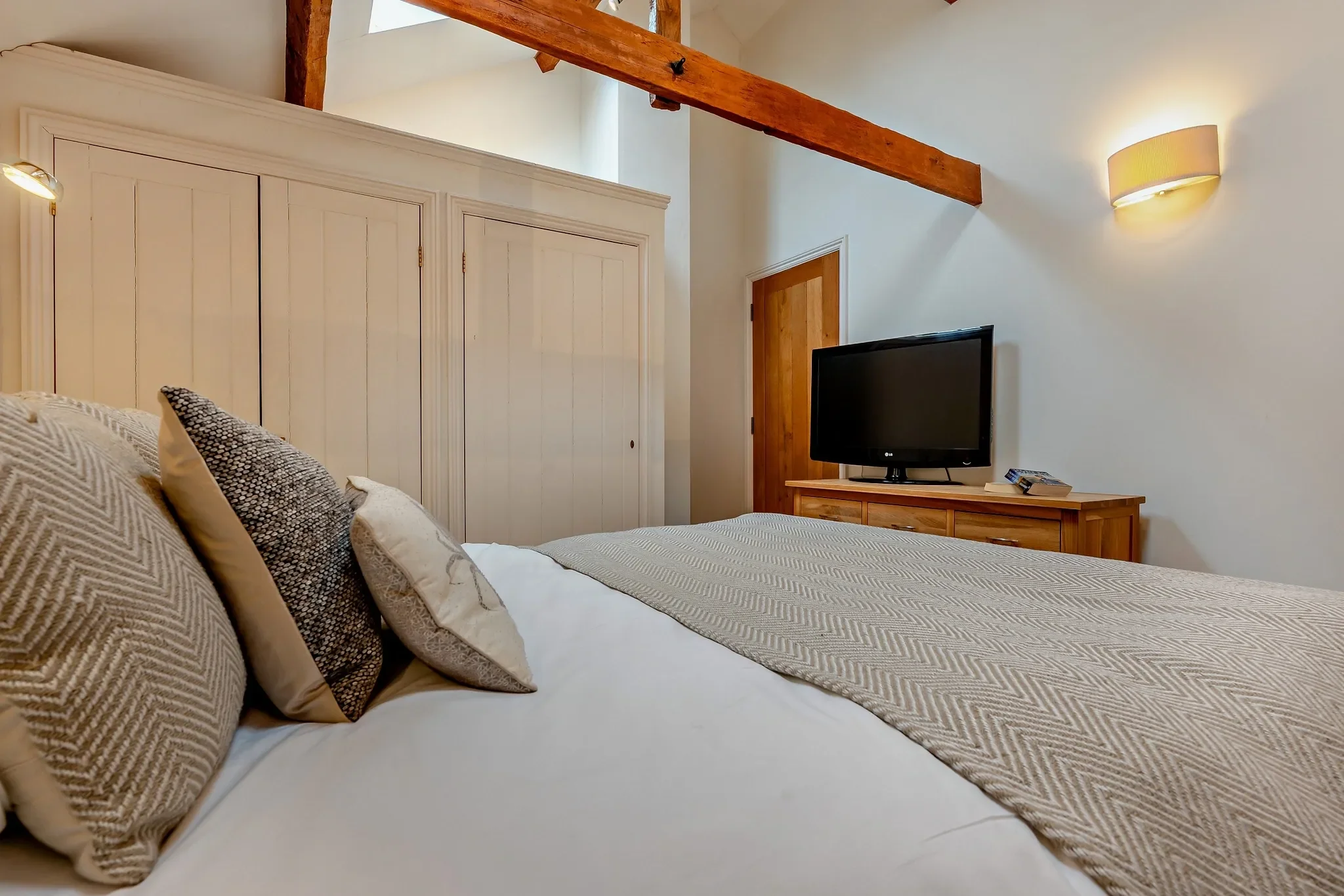 Middle Hill master bedroom from the foot of the bed, highlighting the fitted white wardrobes, TV unit, and exposed roof beams.