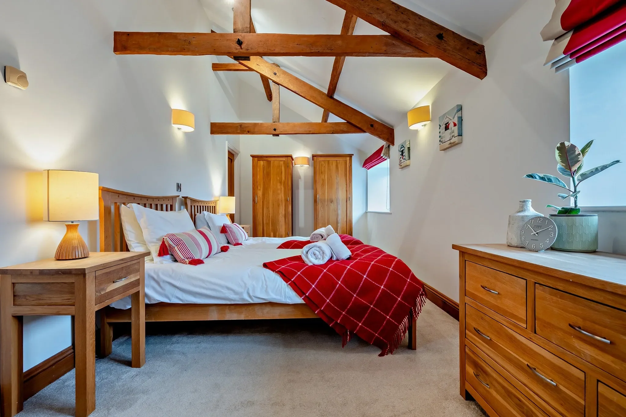 Middle Hill second bedroom configured as a double, with oak furniture, red check throw, vaulted beams, and coastal artwork.