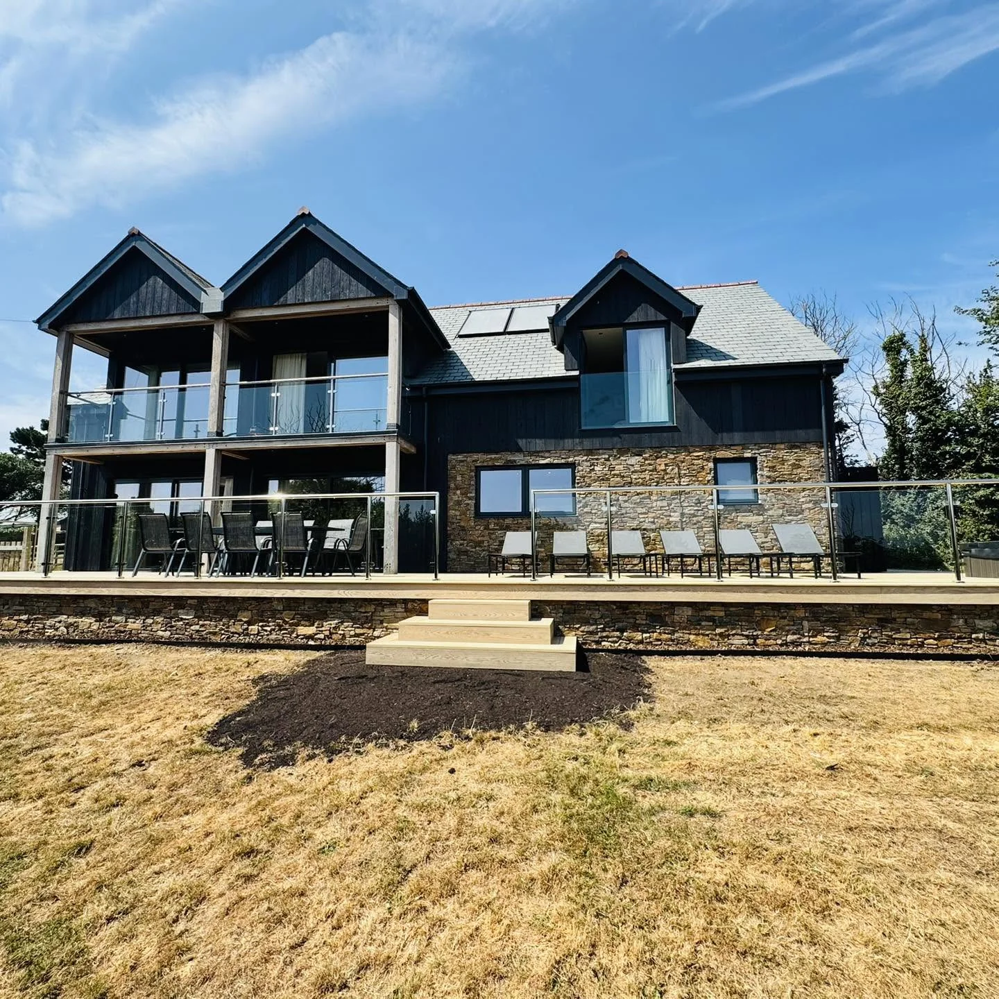 The rear of Nan's House at Whalesborough Farm Resort — black timber and Cornish stone under twin gables, with a raised deck, sun loungers and the private hot tub
