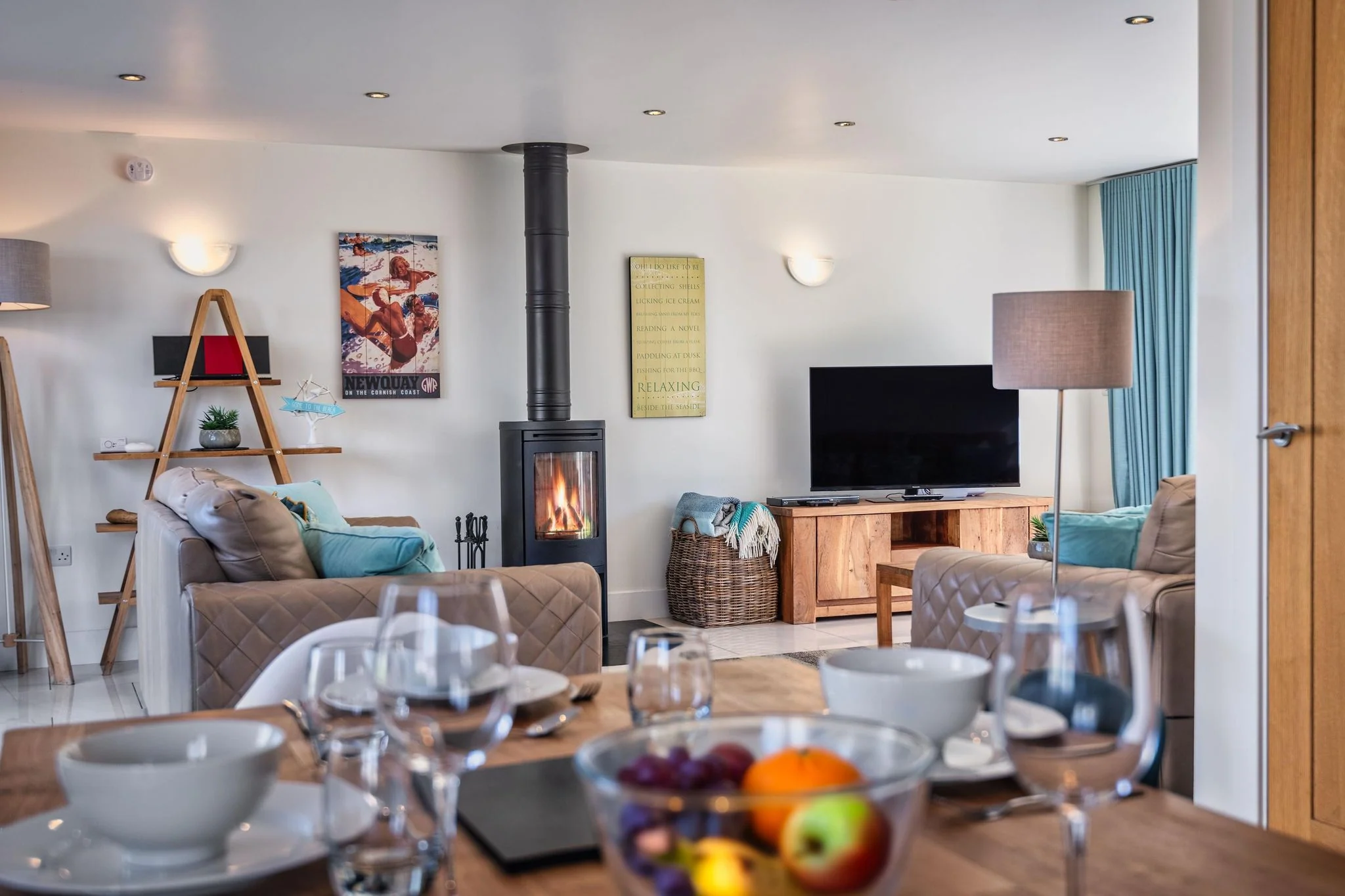 Sheep's House dining table set for six in the foreground, with the lounge, log burner, and TV visible beyond in the open-plan living space.