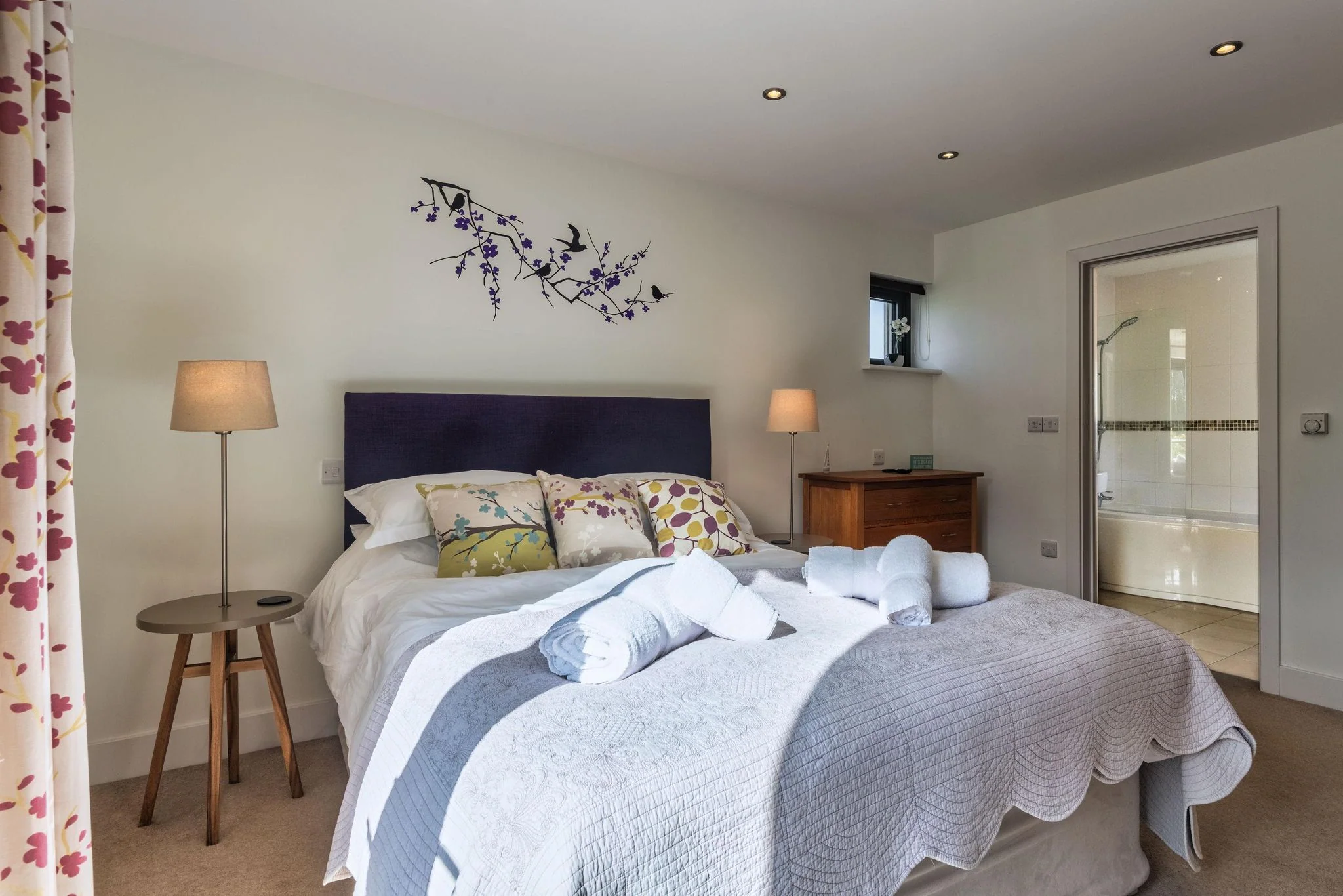 Sheep's House double bedroom with navy headboard, floral curtains, bedside lamps, and an en-suite shower room visible through the open doorway.