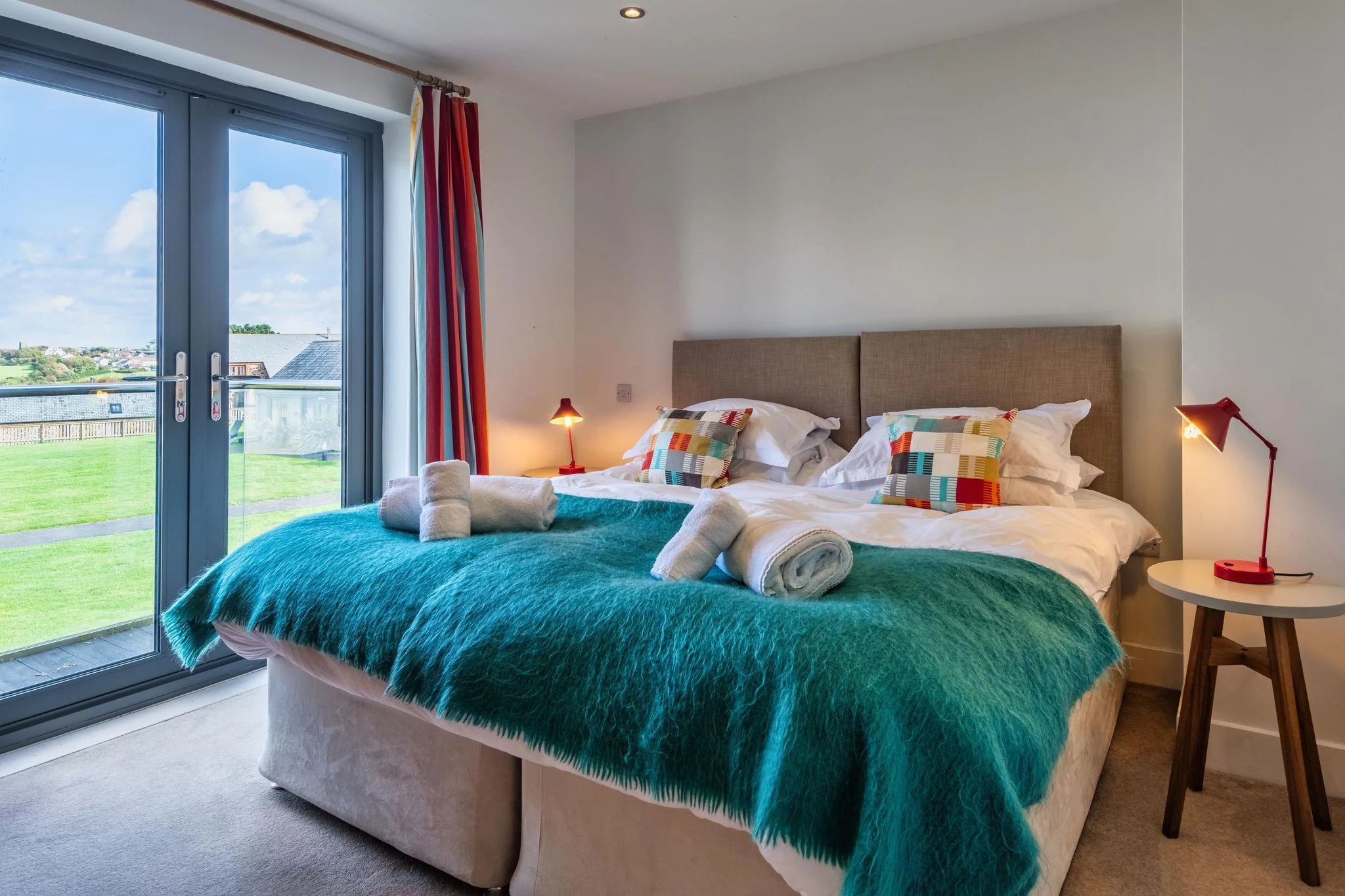 Sheep's House king bedroom with a teal throw, colourful patchwork cushions, and French doors leading onto a Juliet balcony with open countryside views.
