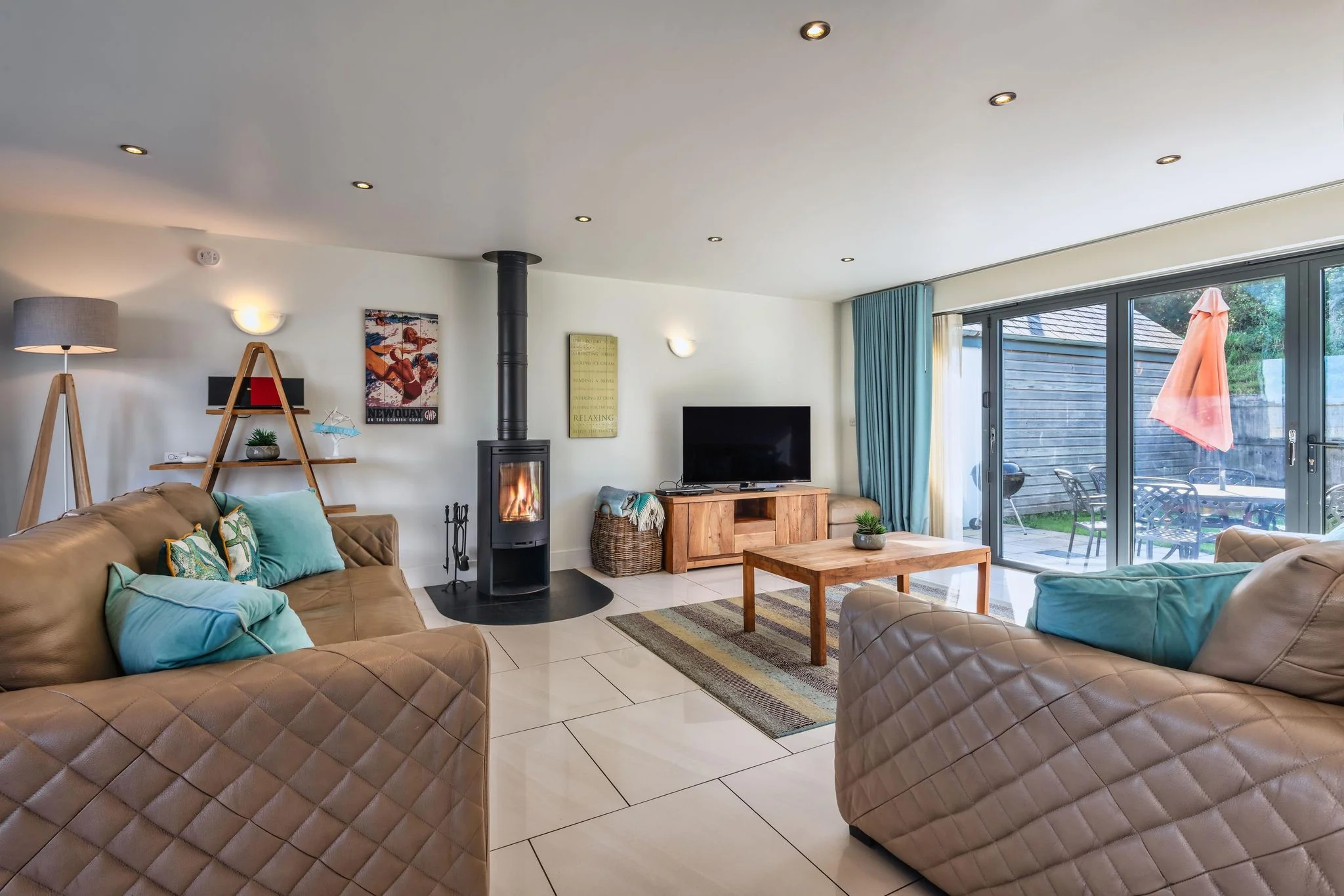 Sheep's House sitting room with quilted leather sofas, lit log burner centrepiece, flat-screen TV, and garden views through the bi-fold doors.