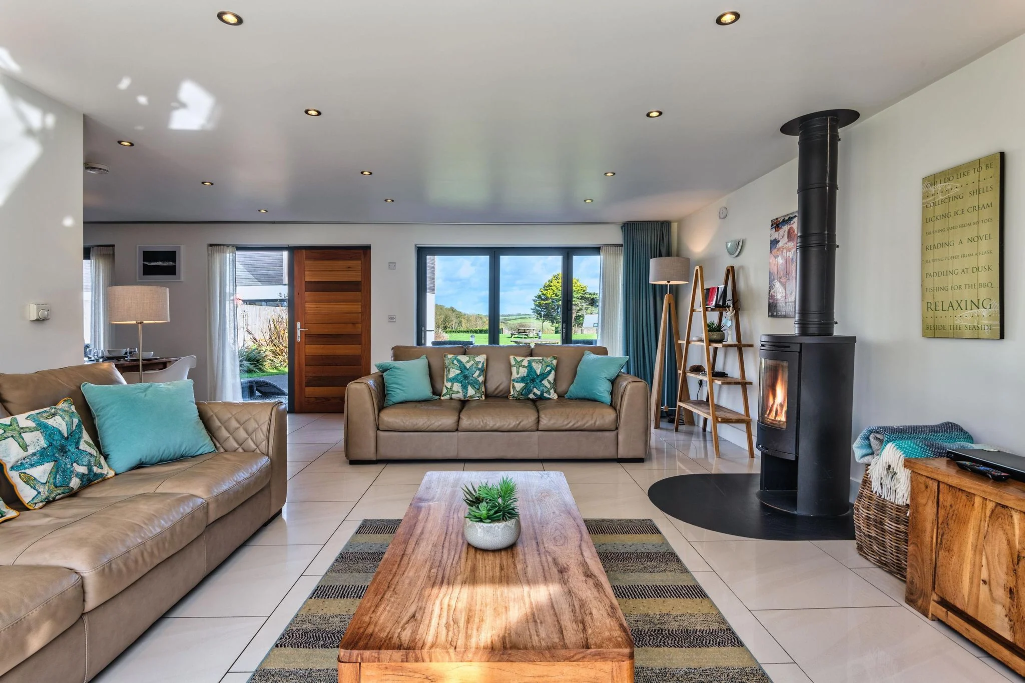 Sheep's House spacious lounge from the far end, showing the full sofa arrangement, log burner, wood-burning stove, and countryside window outlook.