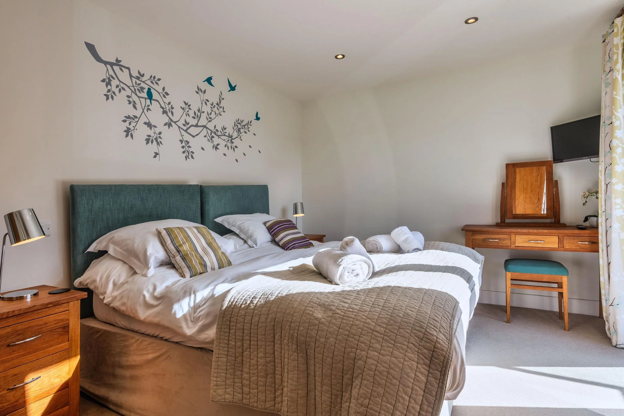 Sheep's House master bedroom with a teal upholstered headboard, bird-branch wall art, dressing table, and flat-screen TV.