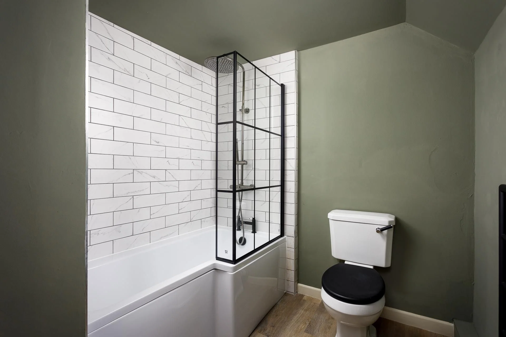 The Farmhouse bathroom with a freestanding bath, overhead rain shower behind black steel-framed glass panels, and sage-green walls.