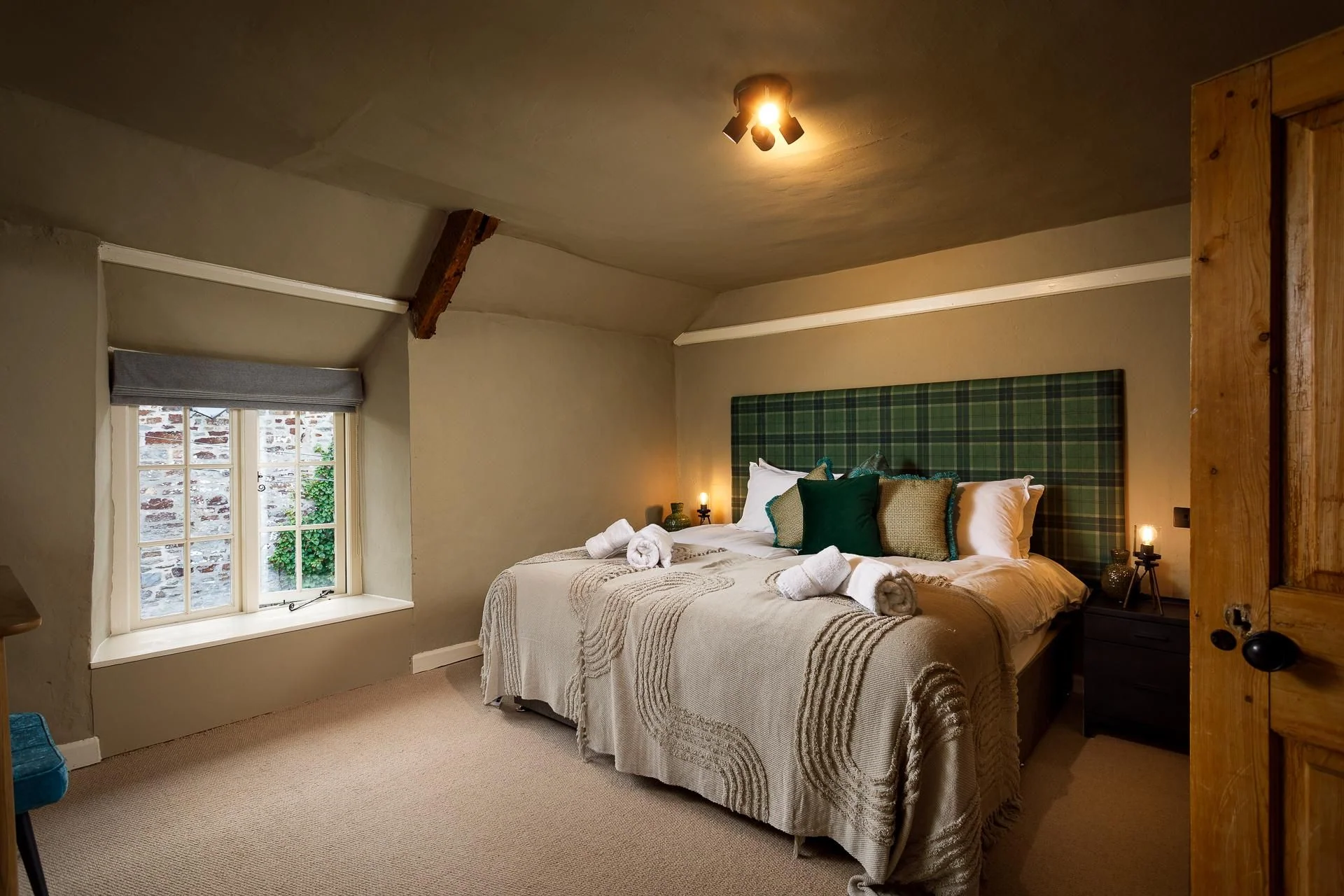 The Farmhouse double bedroom with a tartan-upholstered headboard, warm bedside lighting, and exposed stone walls beyond the window.