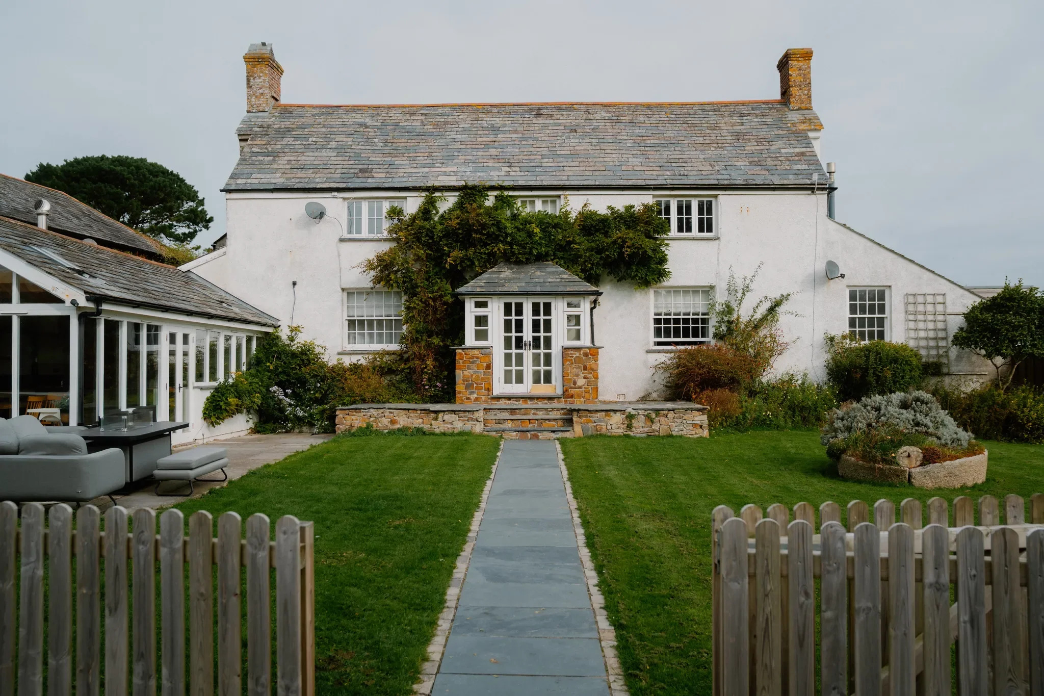The Farmhouse seen from the garden path, a whitewashed Cornish farmhouse with slate roof, climbing plants, and manicured lawn.
