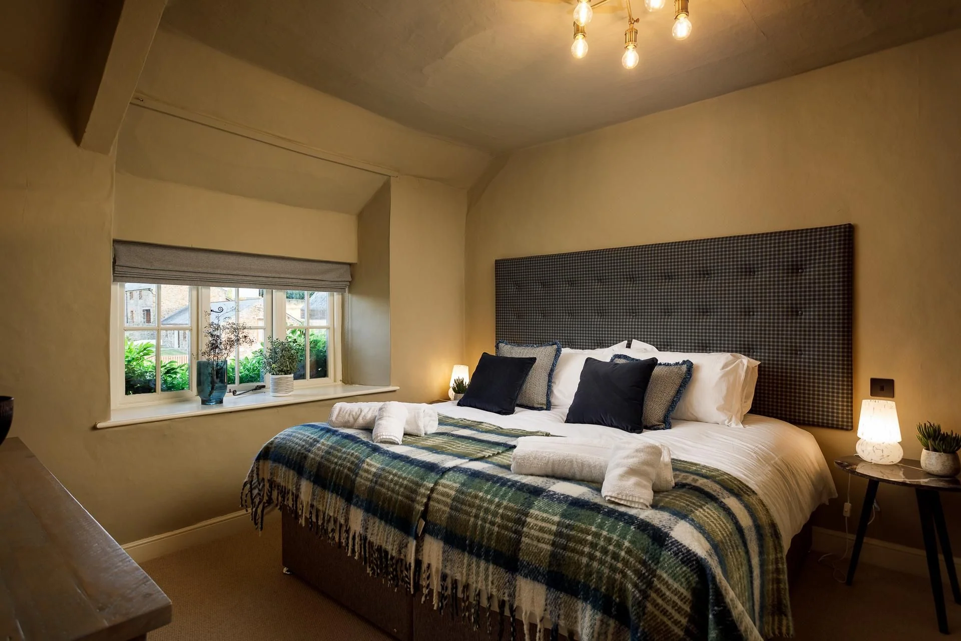 The Farmhouse master bedroom with a tufted dark headboard, tartan throw, and soft pendant lighting above the double bed.