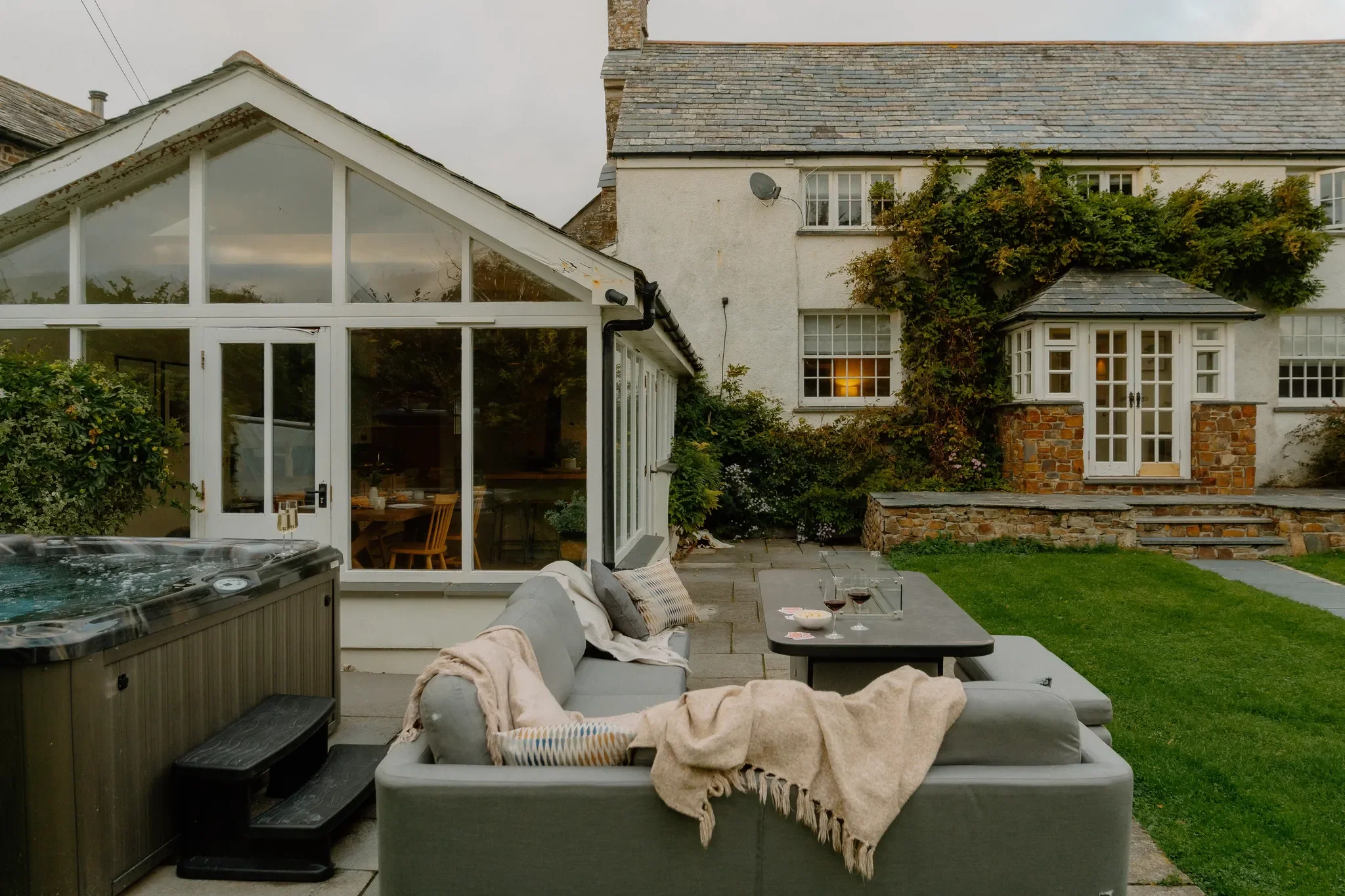 The Farmhouse rear terrace with outdoor sofas, a hot tub, and a dusk view of the conservatory and ivy-covered facade.