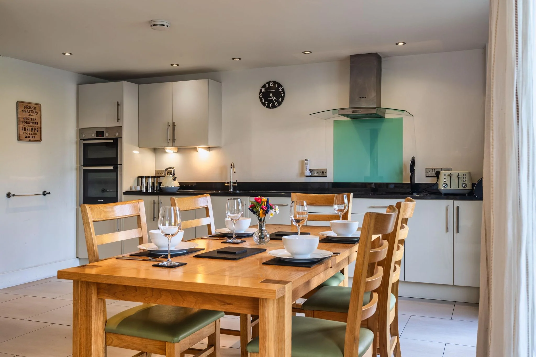 Trevelyan well-equipped kitchen with double oven, green glass splashback and oak dining table set for six.