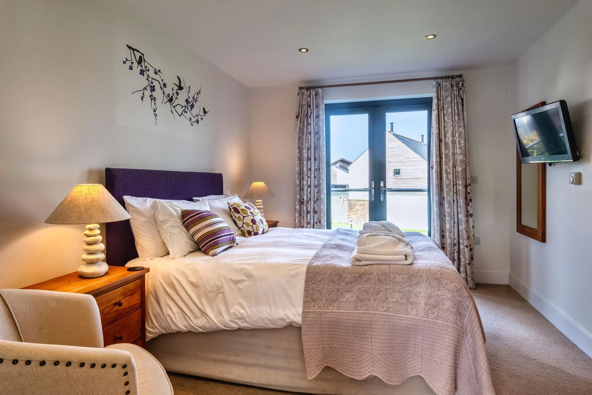 Venners master double bedroom with purple headboard, floral curtains, French doors and wall-mounted TV.