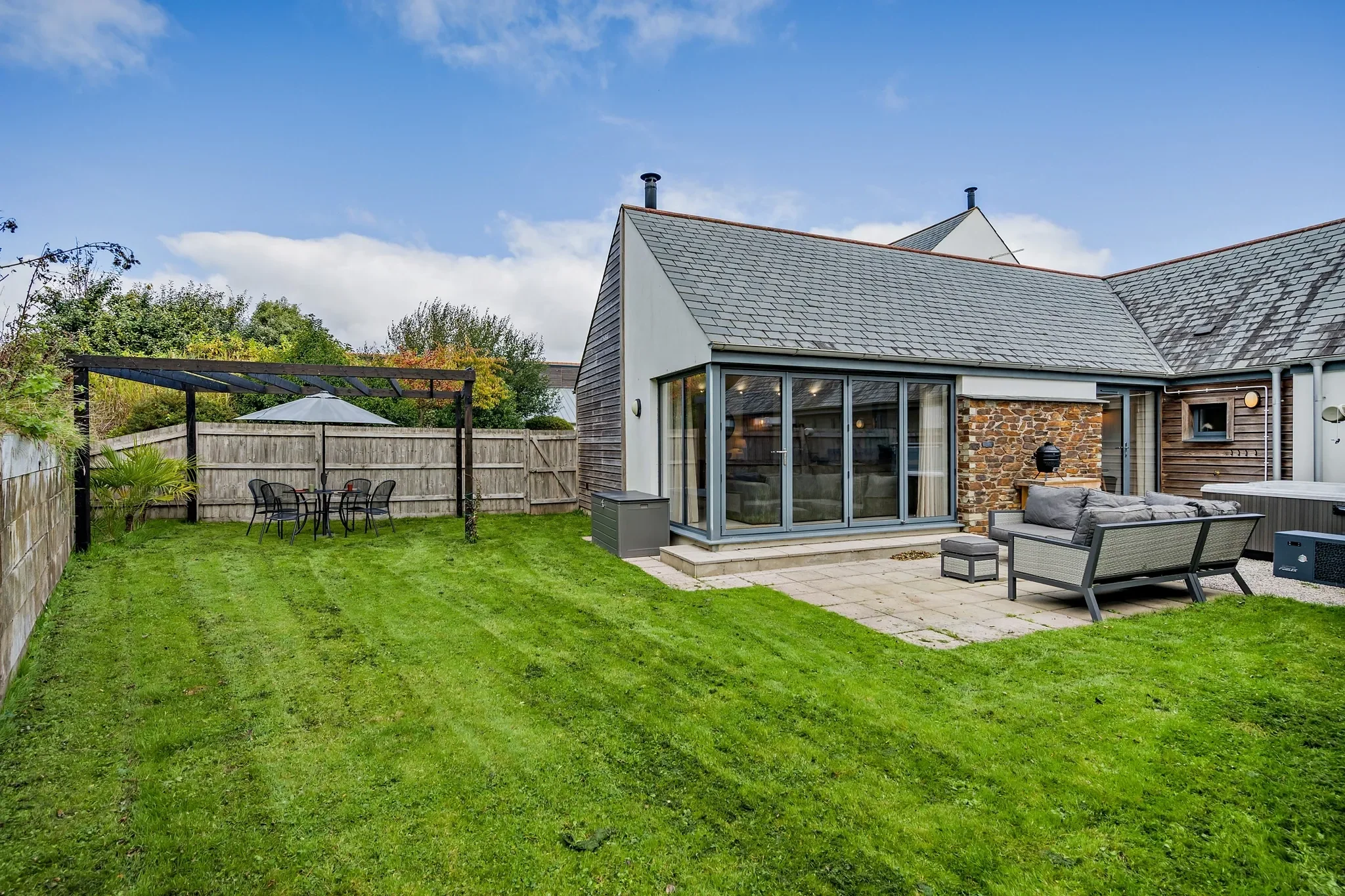 Warrens cottage exterior showing slate roof, stone and timber cladding, glazed bi-fold doors, lawned garden and pergola dining area.