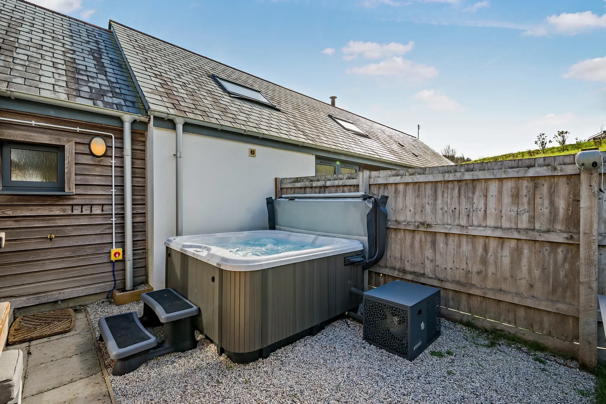 Warrens hot tub on a gravel patio beside the cottage's slate roof and timber cladding under a blue sky.