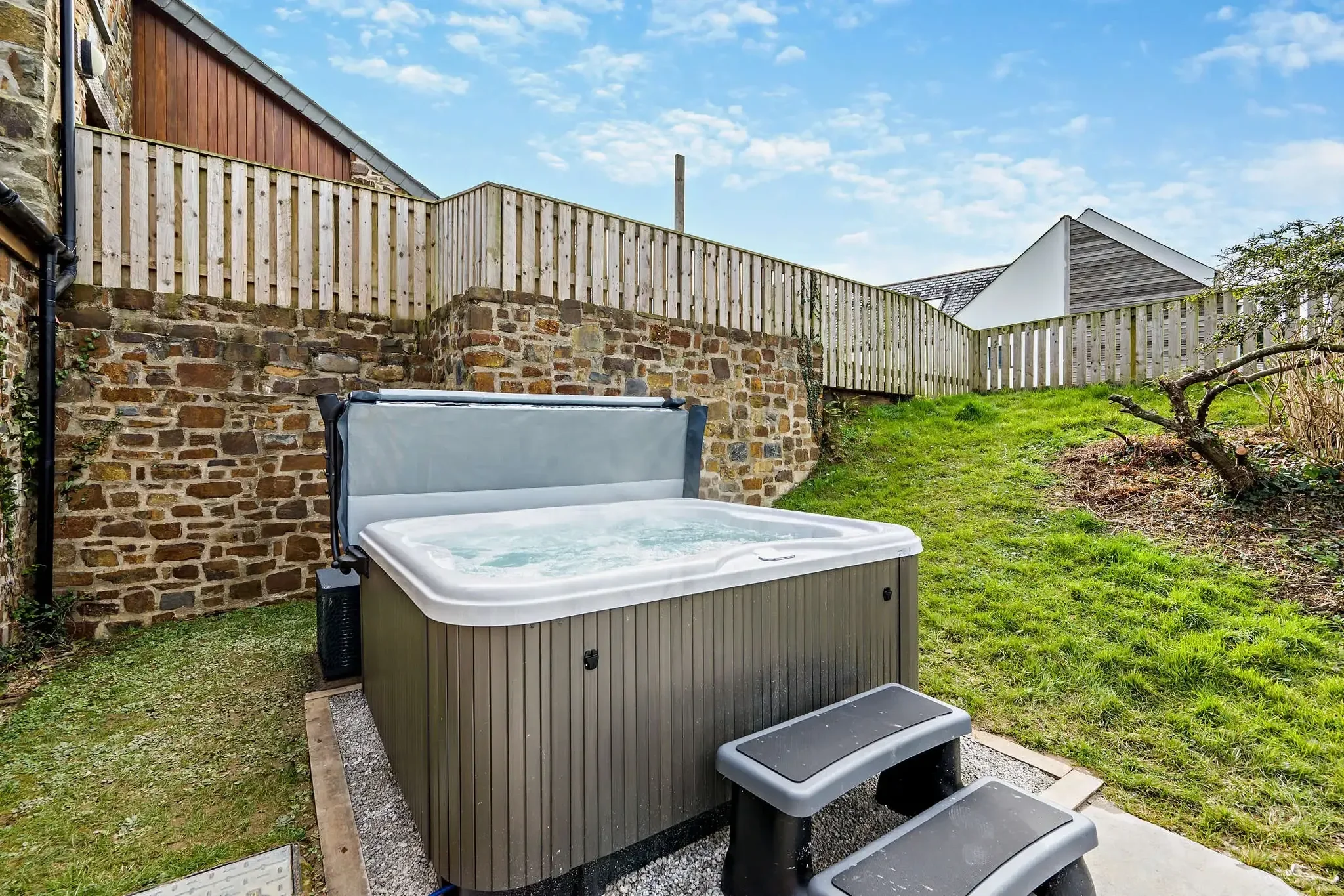 Warrens private hot tub set against a stone wall in the enclosed rear garden with lawned bank behind.