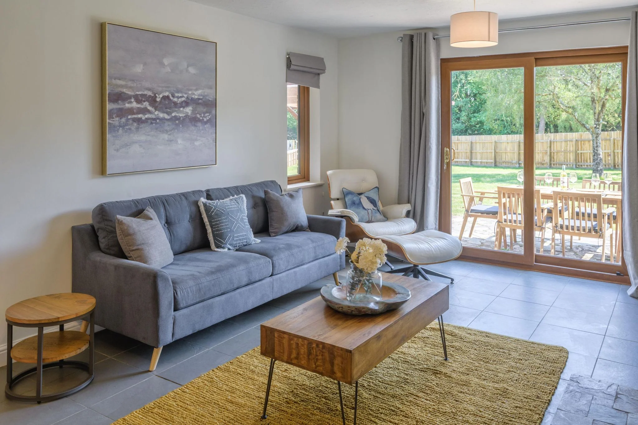 Windy Hills lounge from the far corner, highlighting the blue-grey sofa, mustard rug, and garden views through French doors.
