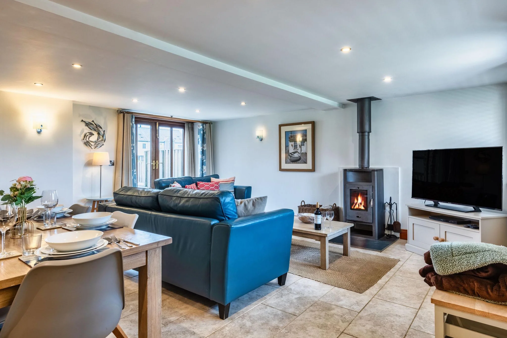 Woody Platt sitting room showing lit log burner, blue leather sofas and flatscreen TV with dining table in the foreground.