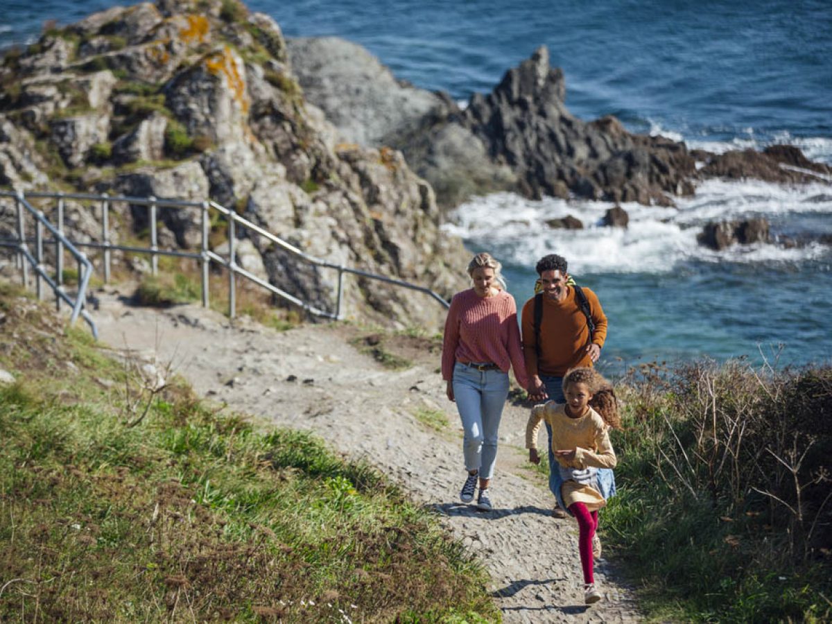 A couple walking up a rock footpath along the cliff edge of Polperro, Cornwall, beside the sea. Their young daughter is running ahead in front of them.